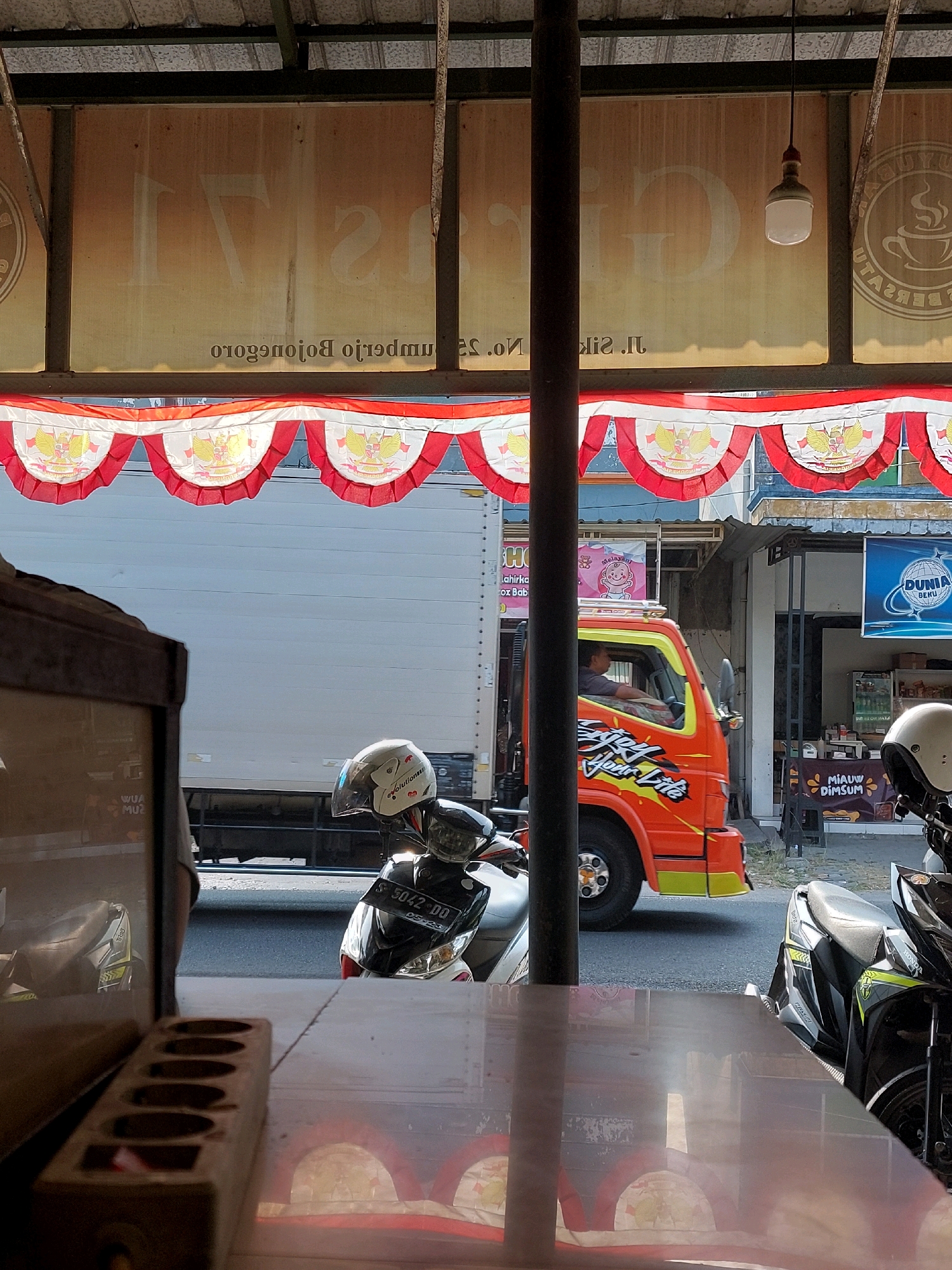This image depicts an outdoor scene in Sumuragung, Indonesia, likely taken from a covered area overlooking a street. In the foreground, a reflective surface, possibly a table or counter, shows reflections of arched structures. To the left, a wooden object with multiple oval openings is visible.
Dominating the mid-ground is a red and orange truck with text that reads "Just Your Life" on its side. A person is visible in the driver's seat. Parked in front of the truck are two motorcycles, one white with a helmet on its seat, and another black and green. The license plate on the white motorcycle is partially visible, reading "S 5042 DD".
In the background, a large white truck or trailer is parked. Above the street, a festive banner with red and white pennants featuring the Garuda Pancasila emblem is strung across. Behind the banner, buildings with signs are visible, including one that says "DUNIA BEKU" (Frozen World). The overall environment suggests a commercial or roadside setting.