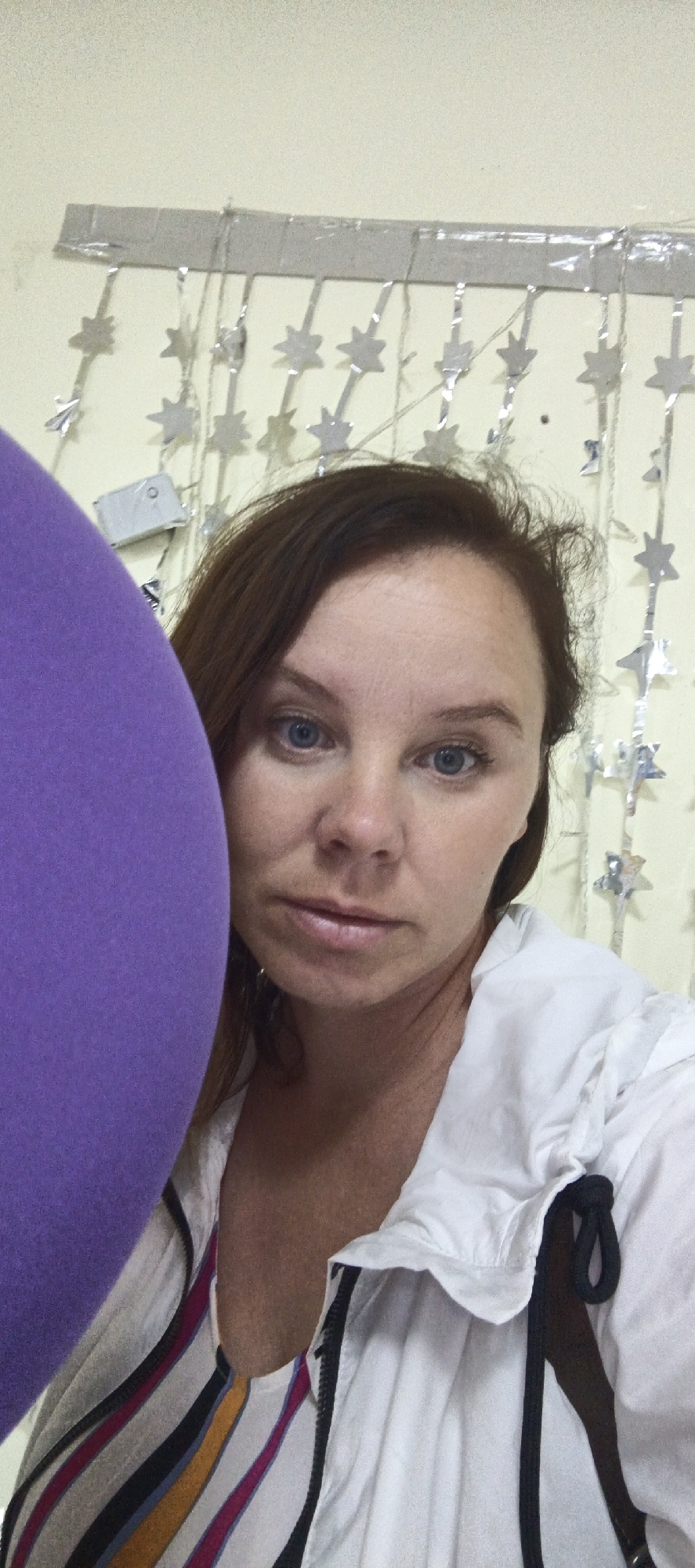 This media file shows a close-up, waist-up selfie of a woman with light skin, brown hair, and blue eyes. She is looking directly at the camera with a neutral expression. A large, purple object is positioned on the left side of the frame, obscuring a portion of her body. She is wearing a white collared shirt with a zippered front. Underneath the white shirt, a colorful striped top with pink, yellow, blue, and white stripes is visible. The background consists of a light-colored wall adorned with a string of silver star-shaped decorations. The overall lighting suggests an indoor setting, possibly daytime. The time of day and weather are not discernible. No visible text is present in the image. The location context of Chaikovsky, Russia, is provided but not visually evident within the image itself.
