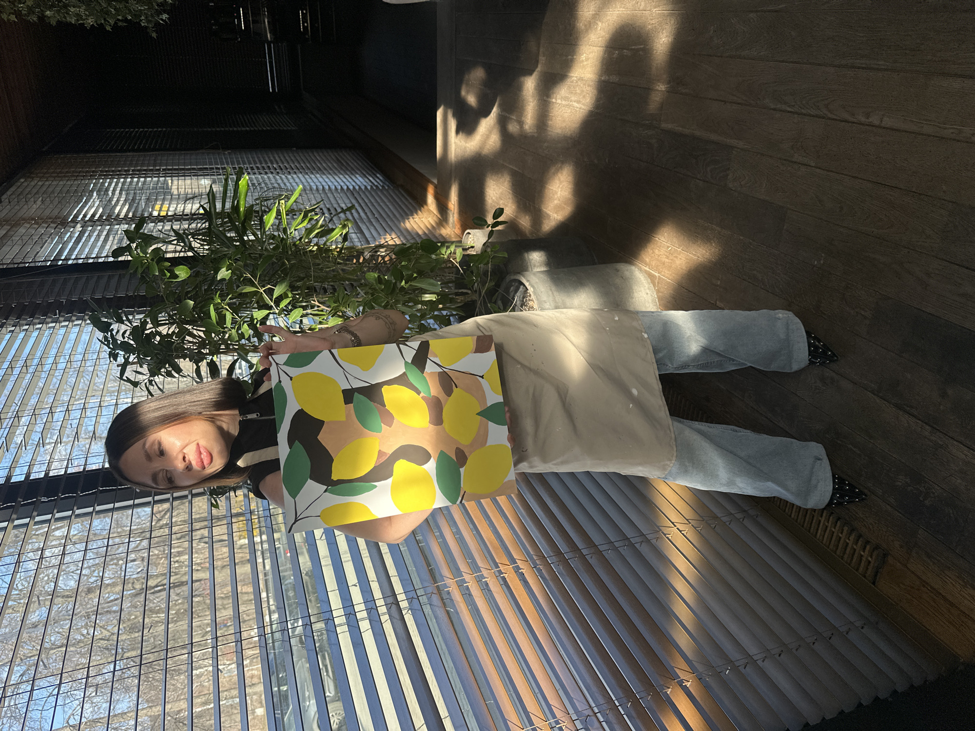 A woman stands inside a room with slatted windows. She holds a lemon-themed painting. She wears blue jeans, black shoes with studs, and a beige apron. The room has wooden floors and a potted plant. The location is in Izhevsk, Russia.