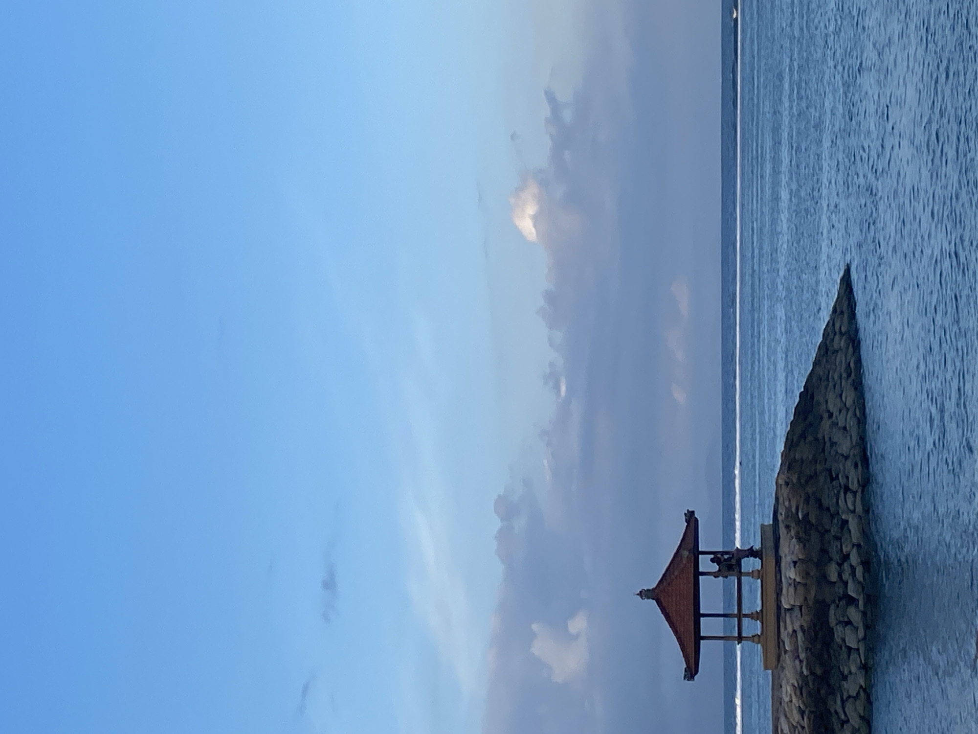 An open-air pavilion with a red, tiered roof sits on a rocky outcrop in the ocean. The pavilion has four columns supporting the roof and appears to be constructed of wood. Two people are visible inside the pavilion. The ocean is a deep blue with gentle waves, and a white line, possibly a boat wake or a distant wave, stretches across the horizon. The sky is a clear blue with scattered, wispy clouds, some of which are tinged with white. The scene is likely the coast of Denpasar, Indonesia, possibly a popular tourist spot or a quiet beachfront area.