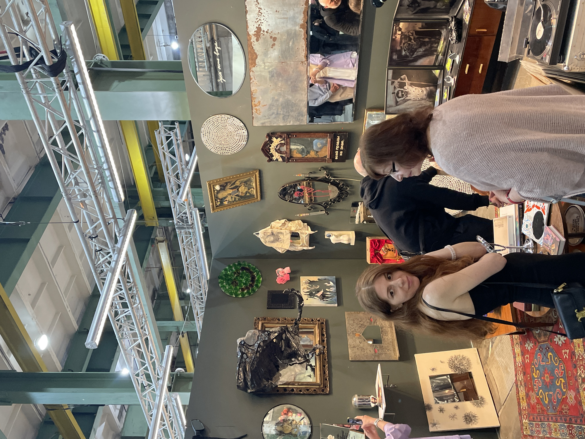 A brightly lit interior space displays a diverse collection of art and artifacts along a dark gray wall, including mirrors, paintings, sculptures, and decorative objects. A woman with long brown hair and wearing a black dress stands in the lower-right, holding a black handbag, and is looking towards the camera. Above her, another woman in a light-colored sweater is examining items on a table. A person in dark clothing is also present, partially obscured behind the woman in the sweater. A red and blue patterned rug is visible at the bottom. In the background, a complex network of metal support beams spans the ceiling, crisscrossed with yellow and green beams. This likely situates the scene within the city of Saint Petersburg, Russia, given the aesthetic and the presence of distinctive artistic and cultural elements.