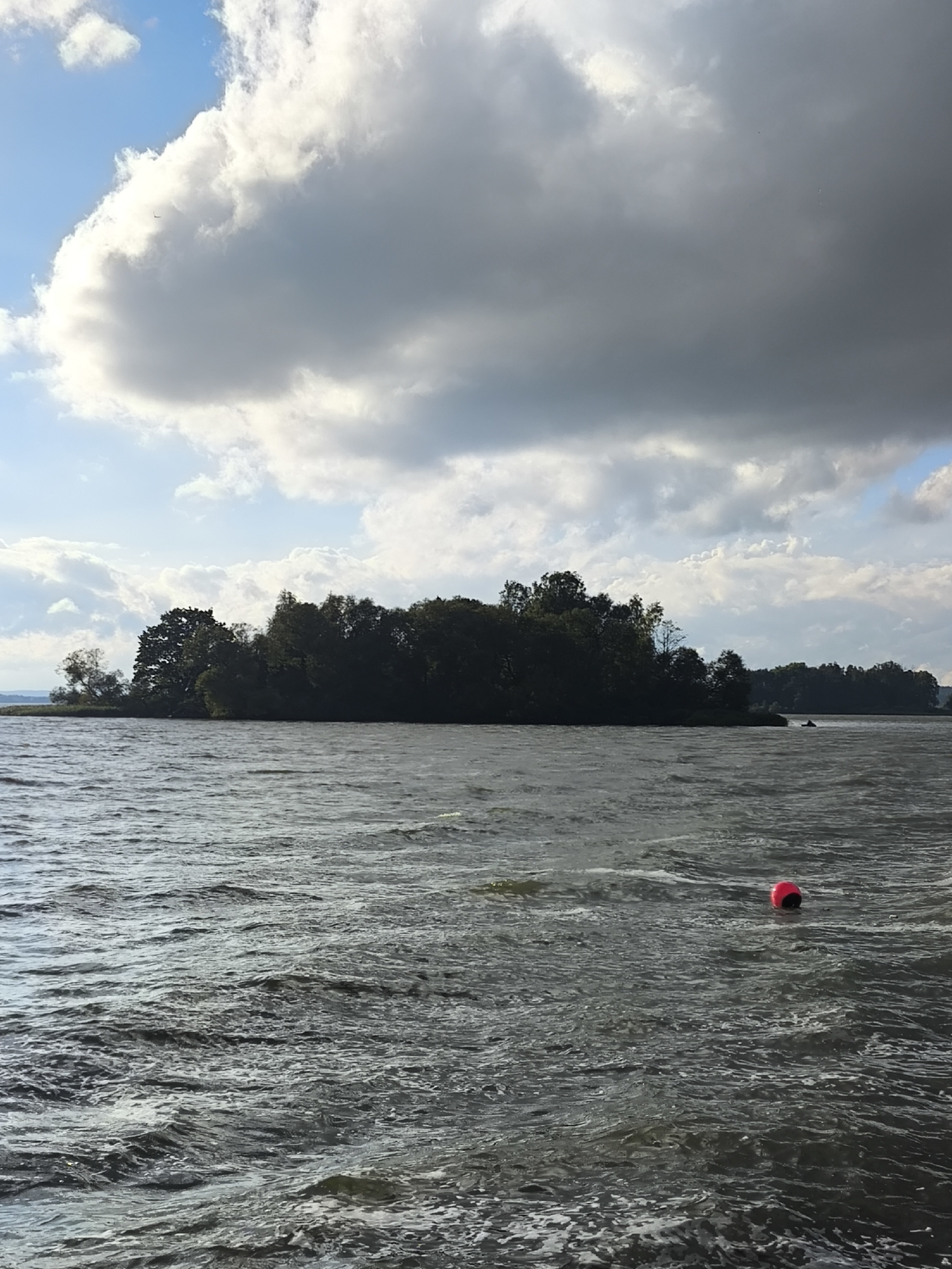 A body of water, likely a lake or river, is depicted under a partly cloudy sky. In the middle distance, a densely wooded island or peninsula stretches across the frame. The water exhibits ripples and waves, suggesting a moderate breeze. In the foreground, to the right, a red buoy floats on the surface. The sky is characterized by a mix of bright blue patches and large, dark, cumulus clouds, with sunlight breaking through in some areas. The overall scene conveys a natural, outdoor environment. The location is specified as Zhdanovichsky Selsoviet, Belarus.