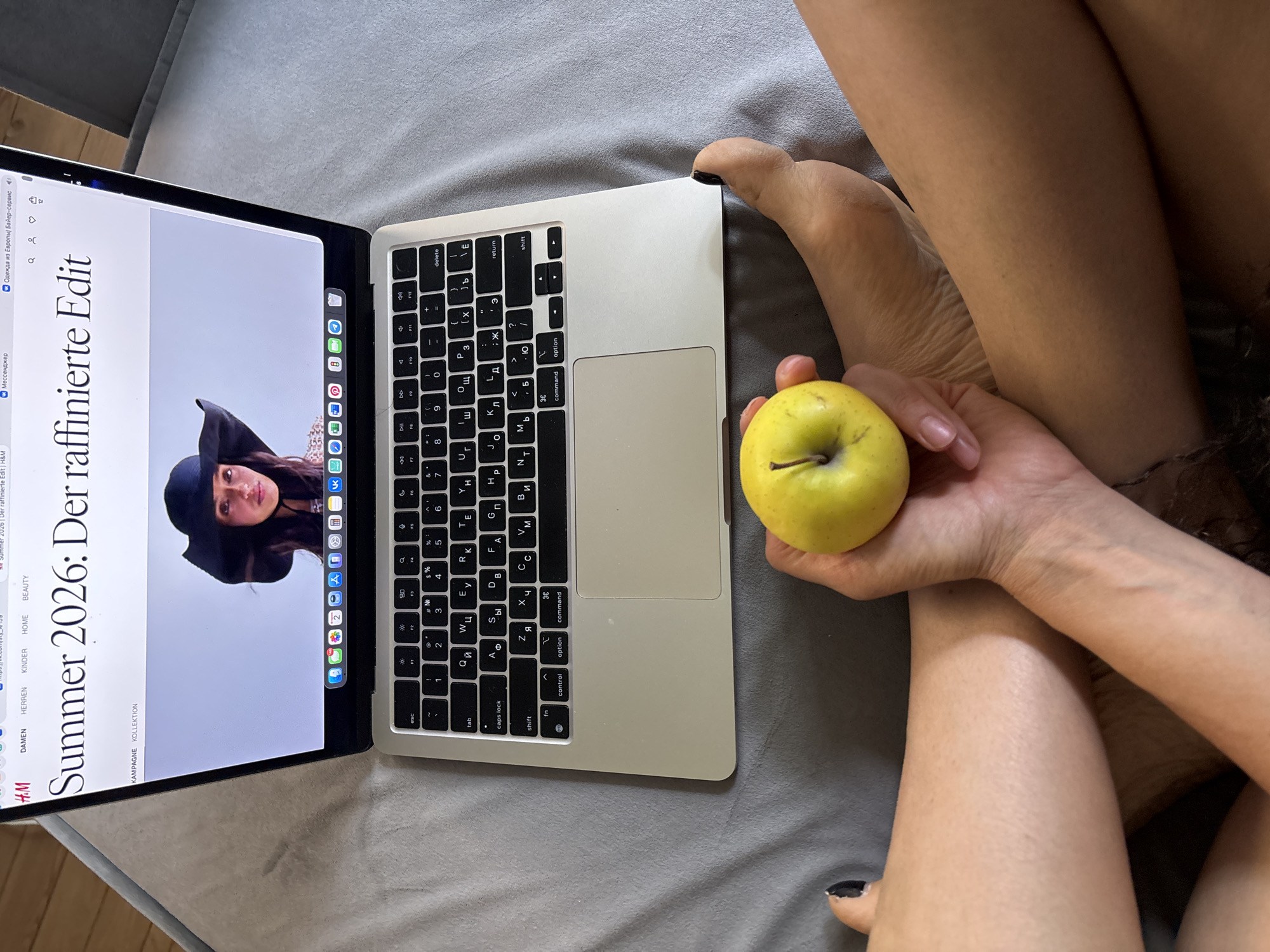 The image captures an indoor scene from a first-person perspective, showing a person's lower body and a hand holding a green-yellow apple, resting on grey bedding or upholstery. A silver MacBook Air laptop is open and positioned on the surface. The laptop screen displays a fashion-related webpage from H&M, featuring the prominent headline "Summer 2026: Der raffinierte Edit" (Summer 2026: The Refined Edit). An image of a woman wearing a wide-brimmed dark hat is visible on the screen.
Other visible text on the screen includes a browser tab labeled "Одежда из Европы Байер-сервис" (Clothing from Europe Buyer-service) and navigation categories such as DAMEN (Women), HERREN (Men), KINDER (Children), HOME, and BEAUTY. The laptop's keyboard has a Cyrillic layout, indicating its use in a Russian-speaking region. One foot is visible, showing a dark nail polish on the big toe. The person is engaged in a relaxed activity, likely browsing online while snacking. The setting is informal, suggesting comfort at home. This scene takes place in Chaikovsky, Russia. No specific time of day or emotions are discernible from the image.