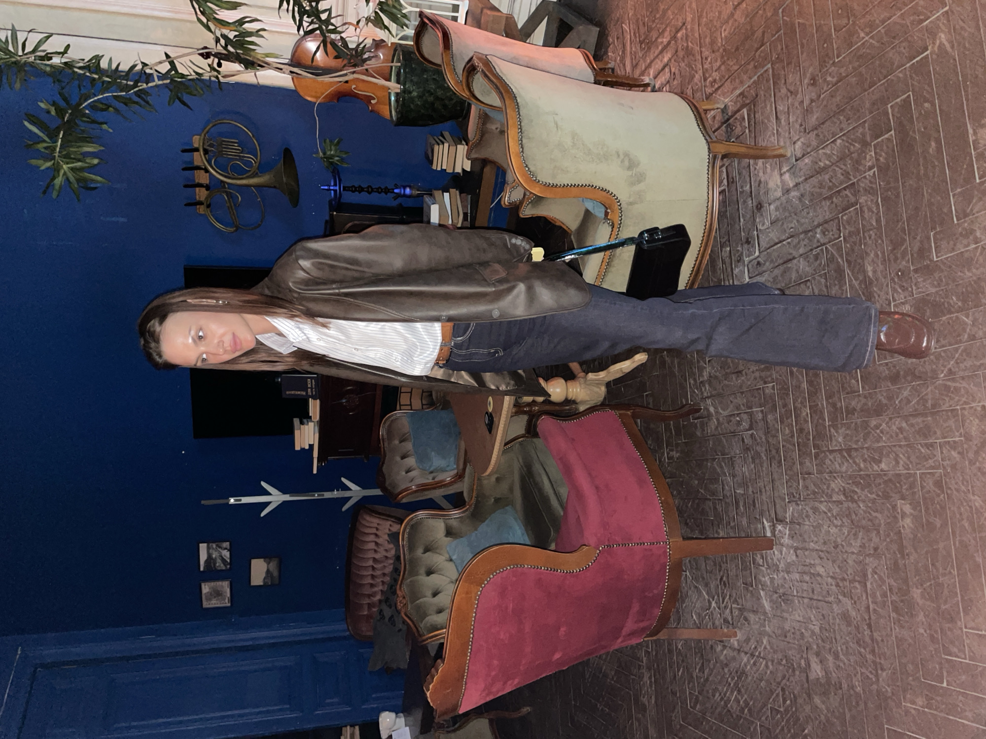 A woman stands indoors among vintage-style furniture. The room is characterized by a dark blue wall on the left adorned with decorations such as a French horn, a bow instrument, and framed images. Vintage chairs upholstered in various fabrics are scattered across a herringbone-patterned floor. The woman wears a brown leather jacket over a striped shirt, dark jeans, and brown shoes. She faces the camera with a neutral expression.
