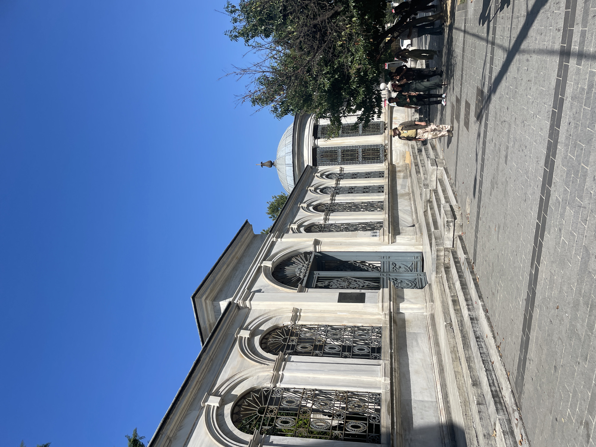 The image shows the exterior of a white marble building with arched windows and a dome. The building is located in Istanbul, Turkey. Several people are walking on the paved walkway in front of the building. The sky is clear and blue. The building appears to be a historic site, possibly a tomb or a mosque. The architectural style suggests it is from the Ottoman era. The presence of a tree on the right side of the image indicates an outdoor setting.