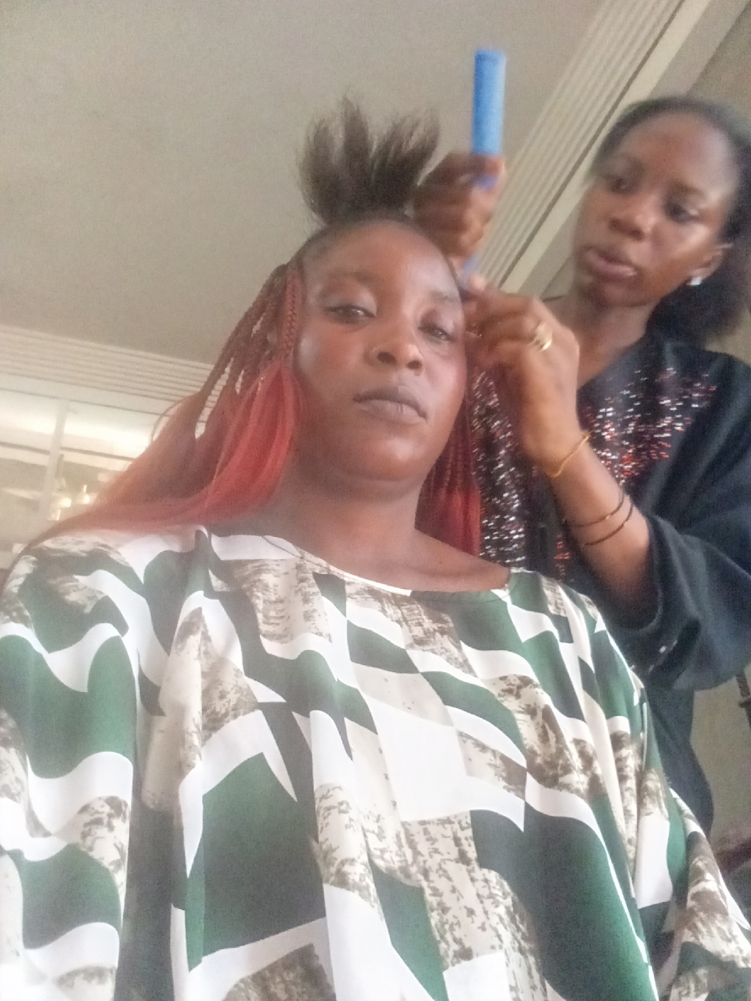 The image features two women indoors, in what appears to be a salon or home setting in Akwanga, Nigeria. In the foreground, a woman faces the camera, her reddish-brown box braids or extensions cascading around her shoulders, with a section of her natural hair pulled up on top of her head. She wears a loose, kaftan-style garment patterned with bold dark green, white, and mottled light brown geometric shapes. Her expression is neutral.
Behind her, a second woman, likely a hairdresser, is actively working on the first woman's hair. She is focused on braiding or styling the hair at the top of the client's head, using a blue hair tool held in her right hand. The hairdresser wears a dark-colored top, possibly black, embellished with sparkling or beaded details on the shoulder. She accessorizes with a gold-colored ring on her right ring finger and several dark bangles on her left wrist. The background shows a light-colored ceiling and what might be part of a window or light fixture, reinforcing the indoor environment. There is no visible text in the image.