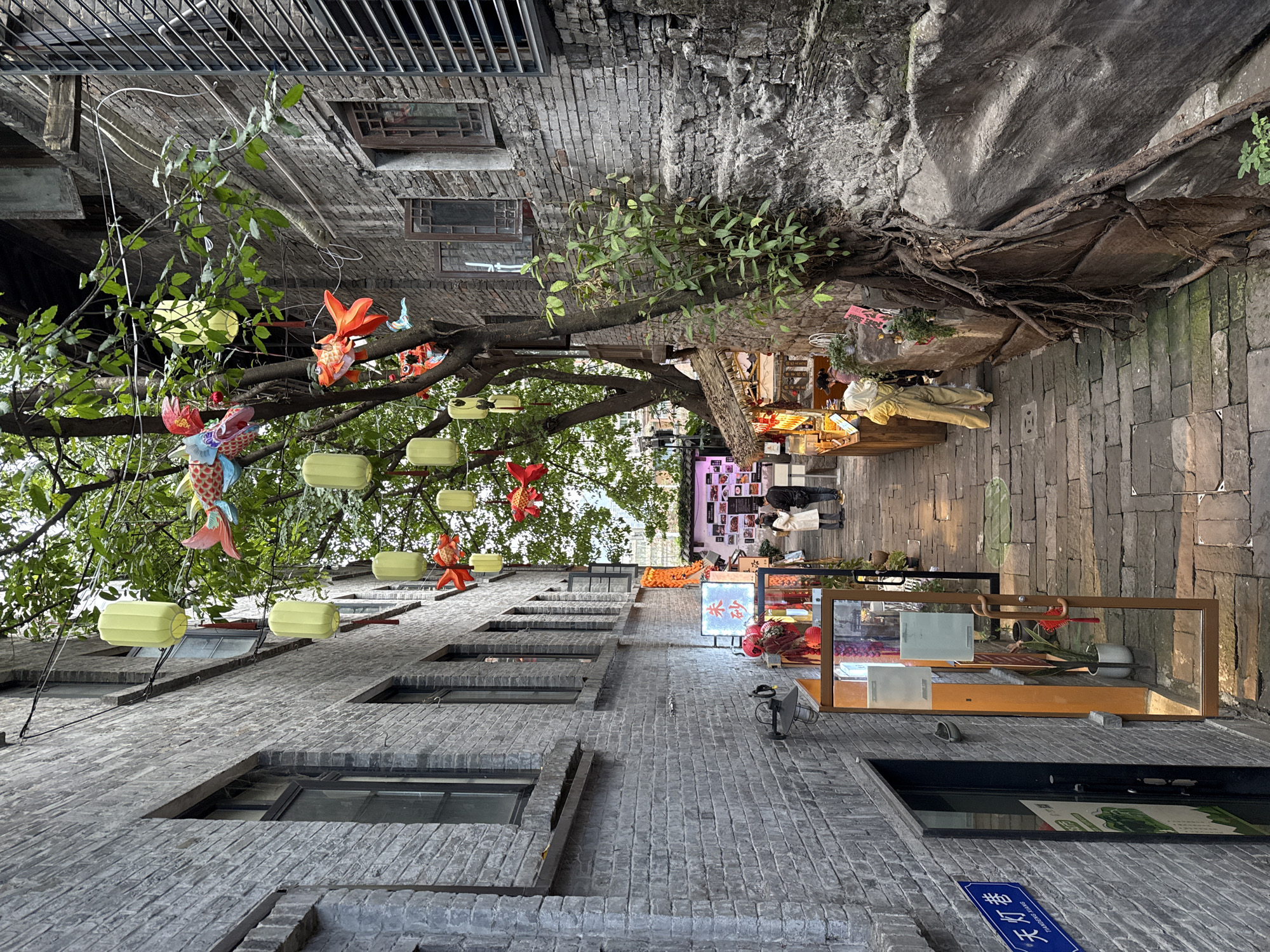 The image depicts a narrow pedestrian alleyway in Yuzhong District, China, identified by a blue street sign on the left wall reading "天灯巷" (Tiāndēng Xiàng). The alley is paved with irregular grey stone blocks.
On both sides, multi-story buildings constructed of weathered grey brick with dark-framed windows line the path. The building on the right features a natural stone or rock wall face at its base, from which large, gnarled tree roots emerge and spread. A tree with green foliage grows within the alley, its branches reaching overhead.
Overhead and among the tree branches, multiple light-yellow cylindrical lanterns are suspended, along with colorful, fish-shaped ornaments in red, blue, and multi-patterned designs. Additional red lanterns and festive decorations are visible near the shop entrances.
On the left side of the alley, a modern shop entrance with a glass door and an illuminated interior is present. On the right, several open-air stalls or small businesses operate. One stall features a thatched roof and various items on display. An illuminated sign with Chinese characters, "朱砂" (Zhūshā), is prominent near one of these stalls. Another display board behind two individuals shows a grid of small images or menu items.
Four individuals are present in the scene. Two adults are standing in the mid-ground, facing towards the right side of the alley, near a display. One wears a light-colored coat, the other a dark jacket. A third person, dressed in yellow overalls and a light head covering, is standing behind a stall on the right, attending to it. A fourth person is partially visible behind another stall on the right.