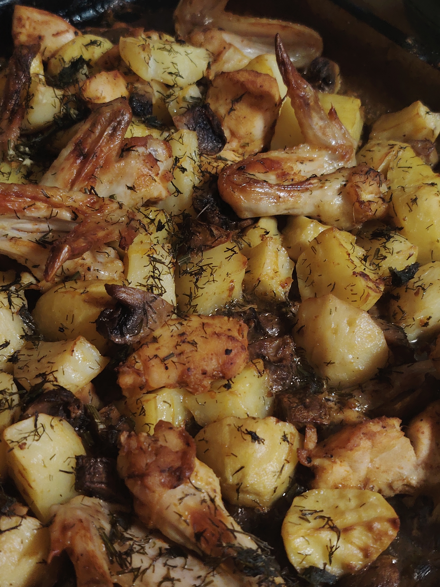 The image is a close-up, top-down view of a dish of roasted chicken wings and potatoes, seasoned with dill and possibly other herbs. The chicken wings are golden brown and appear crispy, with some areas slightly charred. The potatoes are cut into bite-sized pieces and are also roasted to a golden-yellow color, with visible dill sprigs sprinkled throughout. The dish is presented in a dark baking pan, suggesting it was recently cooked. The lighting is warm and highlights the textures of the food. The scene depicts a home-cooked meal.