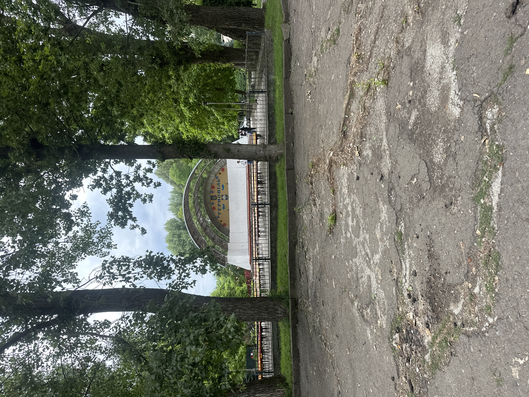 The image shows an outdoor stage in Ternopil, Ukraine. The stage is semi-circular with a white backdrop that features colorful circular elements and text in Ukrainian, which appears to read "ТЕРНОПІЛЬ" (Ternopil). In front of the stage, there are rows of yellow benches. A dark metal fence separates the seating area from a rough, paved ground in the foreground. Several large trees with green foliage frame the scene. In the background, there are more trees and some buildings. Two individuals are visible standing near the stage. The overall environment suggests a public park or open-air venue.