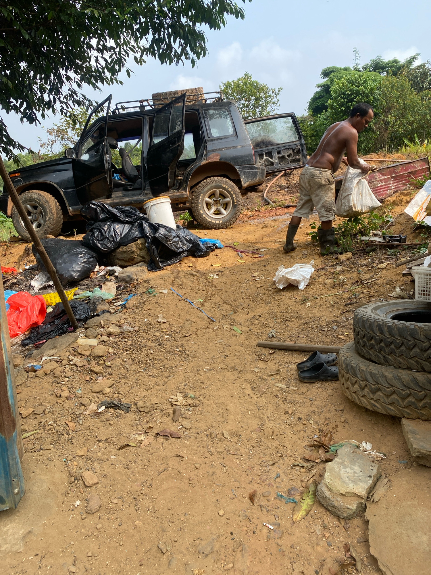 A shirtless man wearing cutoff shorts and dark boots is seen carrying a white plastic bag in a rural outdoor setting. He is standing in a yard littered with trash, including black plastic bags, red plastic bags, and other debris. A black SUV with its doors open is parked nearby, also surrounded by trash. In the background, there are trees and vegetation. The ground is composed of dirt and rocks. Several tires are stacked in the foreground.