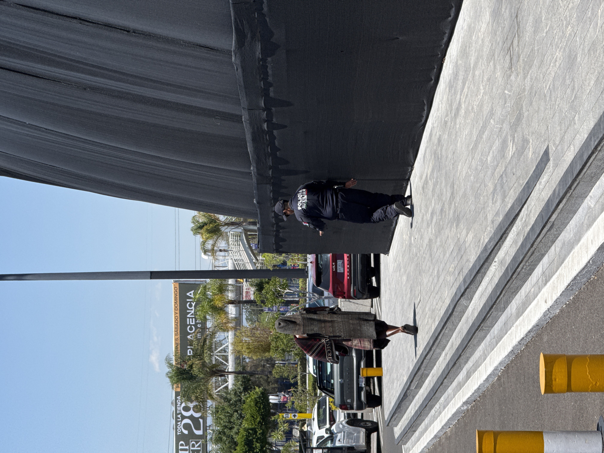 This media file captures an outdoor scene in what appears to be a commercial or public area in Puebla, Mexico, under bright daylight with a clear blue sky. The image is oriented vertically with the sky on the right and the ground on the left when viewed as intended.
A large, dark grey or black fabric structure, possibly a shade canopy or temporary covering, dominates the upper portion of the frame, creating a strong contrast with the bright sky and ground. Below this structure, a man in a dark uniform is visible, with "POLICIA AUXILIAR" (Auxiliary Police) clearly printed on his back. He appears to be standing or walking beside the structure.
Further in the background and to the left, another person is seen walking, wearing a long, patterned garment resembling a poncho and a plaid scarf. Several parked vehicles are visible behind them, including a red SUV with a partially obscured license plate showing "FP-451-", a dark pickup truck, and a white car.
The ground on the lower portion of the image is a light-colored paved surface, possibly a sidewalk or parking area, featuring distinct grey and white striped markings, suggestive of a pedestrian crossing or designated zone. Bright yellow and grey cylindrical bollards line the edge of the paved area on the far left.
In the background, beyond the vehicles, there are palm trees and other vegetation, along with building structures and a prominent billboard. The billboard displays text: "PLACENCIA muebles" (Placencia furniture), "TODA LA TIENDA" (All the store), and "VIERNES, SÁBADO Y DOMINGO" (Friday, Saturday and Sunday), along with the number "28", likely indicating a promotion or sale for a furniture store. The scene suggests a busy commercial environment.