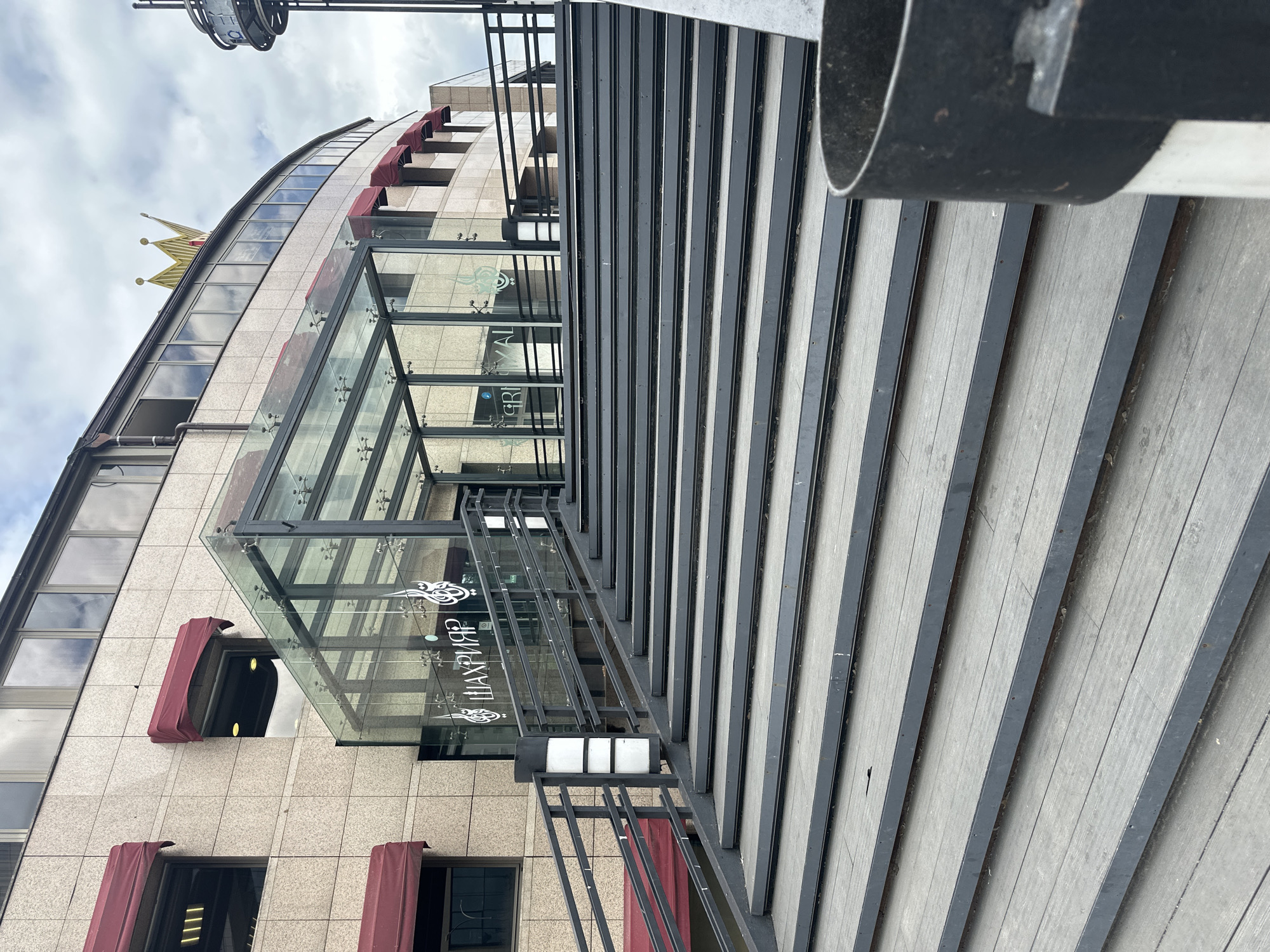 The image shows an outdoor view of the entrance to a building in Minsk, Belarus, featuring a wide set of outdoor stairs leading up to a glass-covered entrance. The building has a textured light-colored facade with dark windows and red awnings. A golden crown sculpture is visible on the roof of the building. Arabic calligraphy is displayed on the glass entrance. The stairs are made of gray wooden planks with dark metal edging. A black metal railing runs along the sides of the stairs, with a lamp post on the left side. The sky is overcast. There are no people visible in the image.