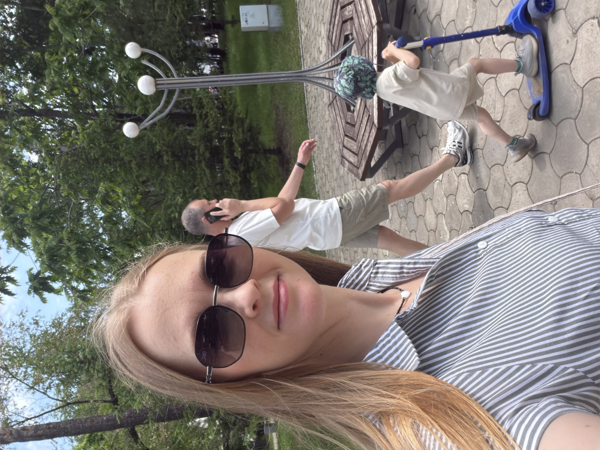 The image shows a woman taking a selfie in a park. She is wearing sunglasses and a striped shirt. In the background, a man is talking on his phone and a child is riding a scooter. The park has trees, a lamp post, and a wooden bench. The location is identified as the city of Khabarovsk, Russia. The pavement is made of interlocking concrete pavers. The scene appears to be a pleasant, outdoor setting during the day.