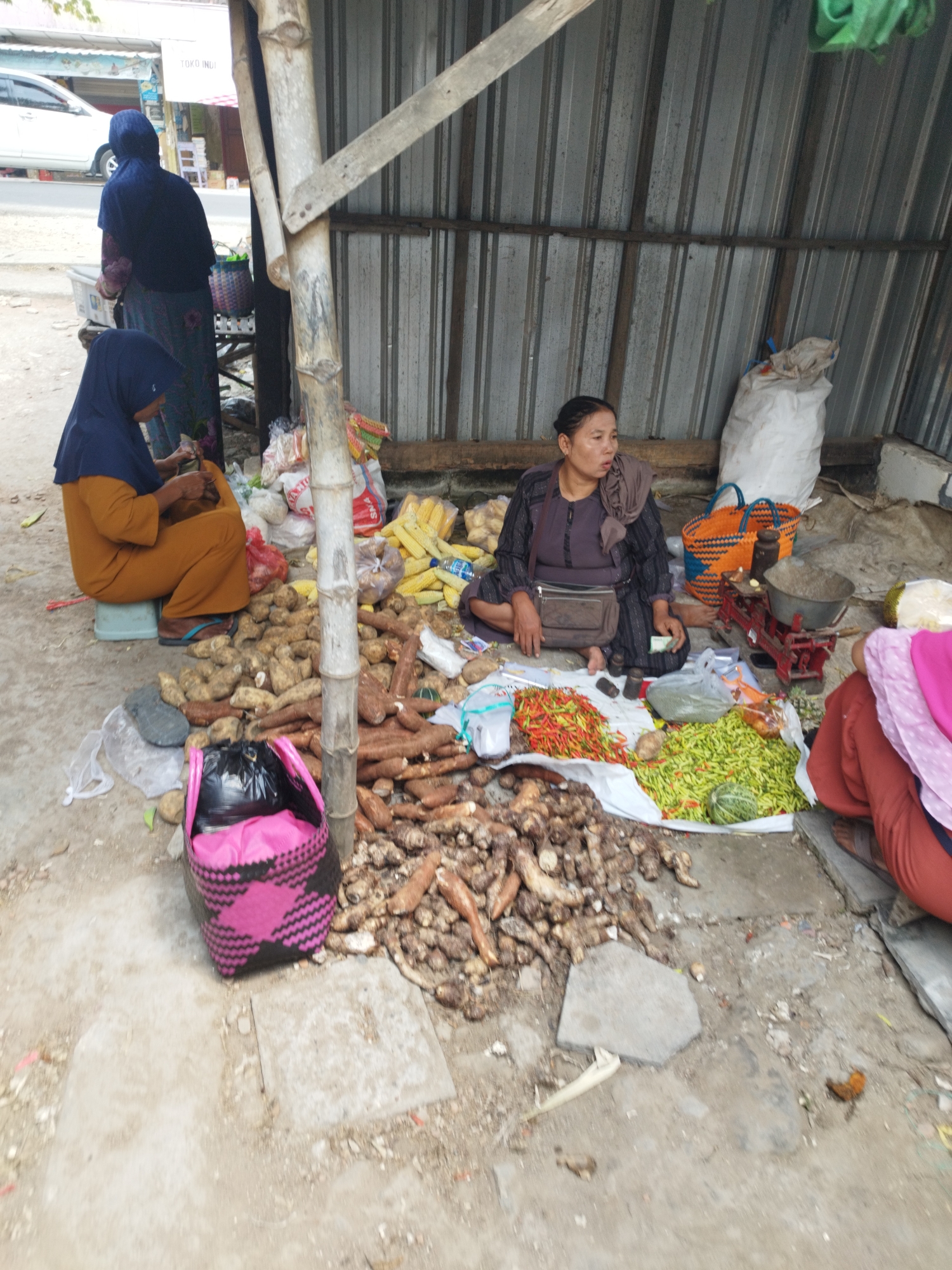 In Bumirejo, Indonesia, an outdoor market stall features a woman sitting on the ground, surrounded by produce. She is wearing a dark patterned dress and a headscarf, with a small crossbody bag. In front of her, arranged on a white tarp, are piles of chili peppers, both red and green, and other vegetables. Behind her, a large orange and blue woven basket sits next to a red antique scale. Further back, a white sack is visible, leaning against a corrugated metal wall that forms the roof of the stall. To her left, another woman in an orange tunic and dark blue headscarf is seated on a stool, attending to something in her hands, with a black and pink woven basket next to her. Various root vegetables, possibly cassava and yams, are piled on the ground around the seated women and in front of the stall. A white car is partially visible in the background on a paved road. The ground is a mix of dirt and concrete.