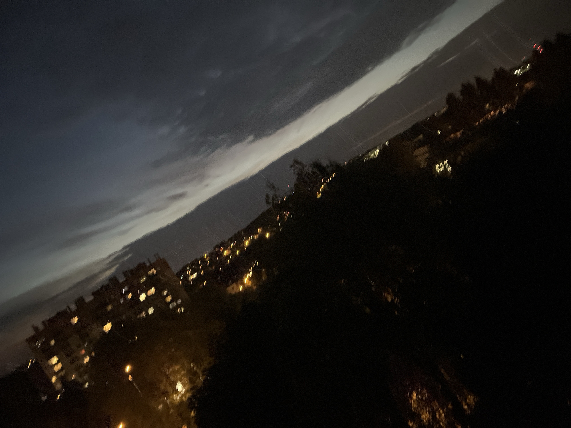 The image is a tilted, dark, and blurry nighttime photograph of a city skyline. The scene is dominated by deep shadows and scattered, out-of-focus lights. In the upper left portion of the image, there are indications of buildings with glowing windows, suggesting an urban environment. These lights are yellowish and appear as soft orbs due to the low light and blurriness.
Below these lights, a dark, silhouetted mass appears to be trees or vegetation. Further down and to the right, more scattered lights can be seen, some appearing as tiny dots and others as streaks, possibly from moving vehicles or distant streetlights.
The sky occupies the right and upper portions of the image. It is dark and cloudy, with a diagonal band of lighter gray or white cloud streaking across it, creating a stark contrast with the dark sky. The overall impression is one of a hazy, possibly overcast night in a city. No people or specific identifiable landmarks are visible due to the darkness and blur.