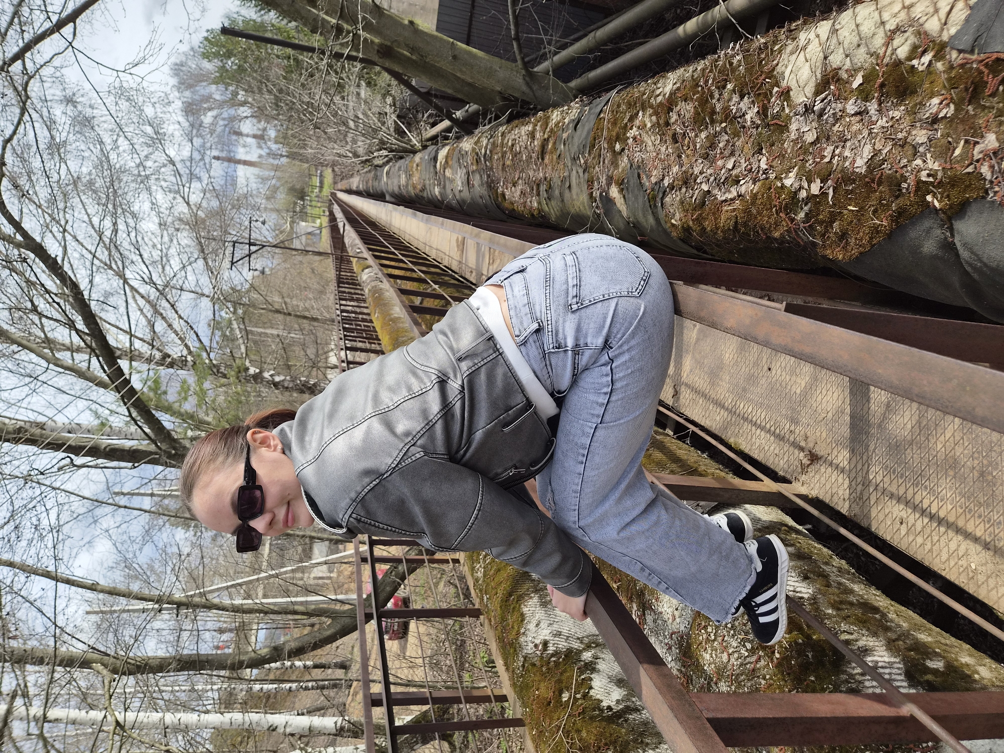 A female stands on a rusted iron grate walkway next to a large pipe covered in moss and debris in the city of Chaikovsky, Russia. She wears blue jeans, a light-grey leather jacket, and black-and-white Adidas Hamburg shoes. She leans forward with her hands on the walkway railing, her face visible, wearing sunglasses. The background includes bare trees and industrial structures under an overcast sky.