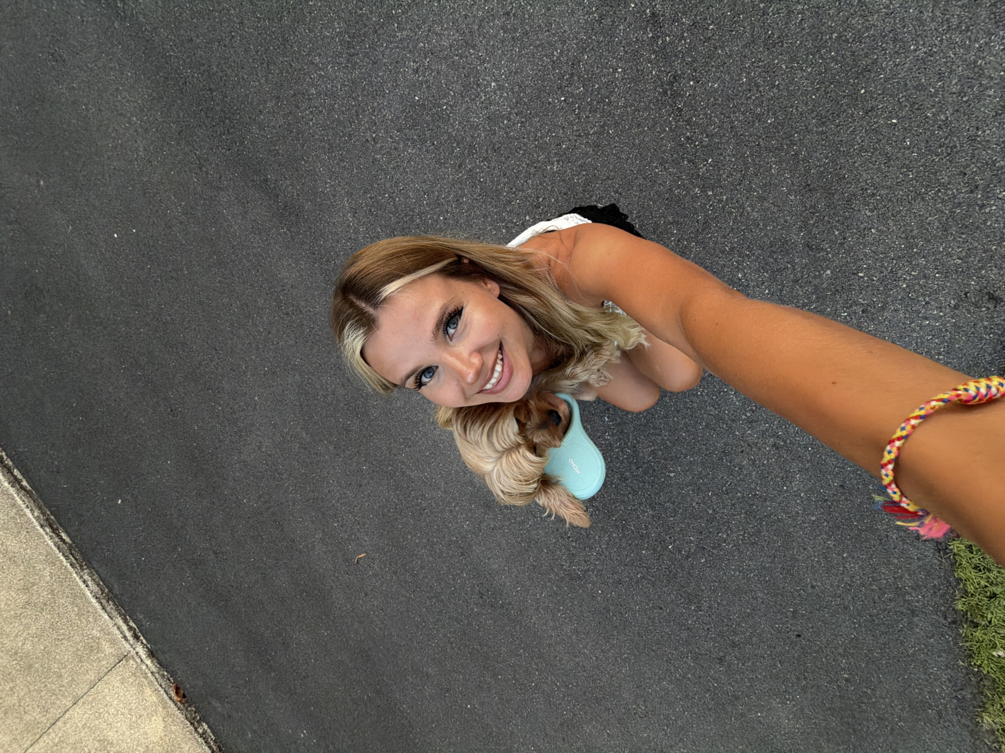 The overhead shot shows a young woman and a small dog on an asphalt surface. The woman, with blonde hair and blue eyes, is smiling and looking up at the camera. She is wearing a white top with black trim. The dog, a small breed with light brown fur, is nestled next to her, with its head resting on a light blue object, possibly a dog toy or leash holder. The woman's right arm is extended towards the camera, and she is wearing a colorful, braided bracelet on her wrist. The asphalt appears to be a road or path, with a lighter colored concrete curb on the left side of the image and a patch of green grass on the right. The lighting suggests it is daytime, likely outdoors. The overall mood is cheerful and casual. No visible text is present in the image.