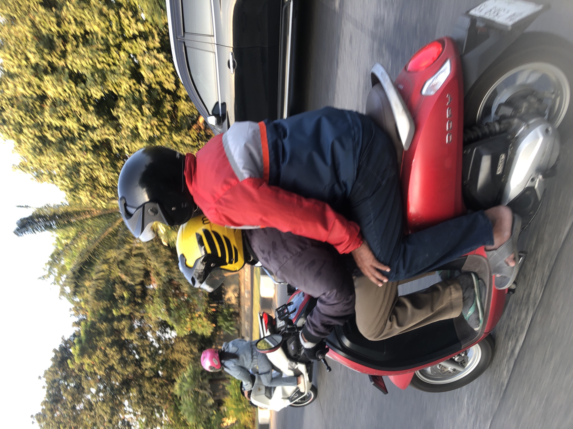 A couple rides a red scooter in the city of Candi, Indonesia. The male driver, wearing a black helmet, is in the front, dressed in a red, gray, and blue jacket, and blue jeans. The female passenger behind him wears a camouflage jacket and brown pants, with a yellow helmet. A white scooter carrying a woman in a pink helmet and denim jacket is visible in the background. A black SUV is to the right of the red scooter. The scene is set outdoors with lush green trees and a bright sky, suggesting it's daytime.