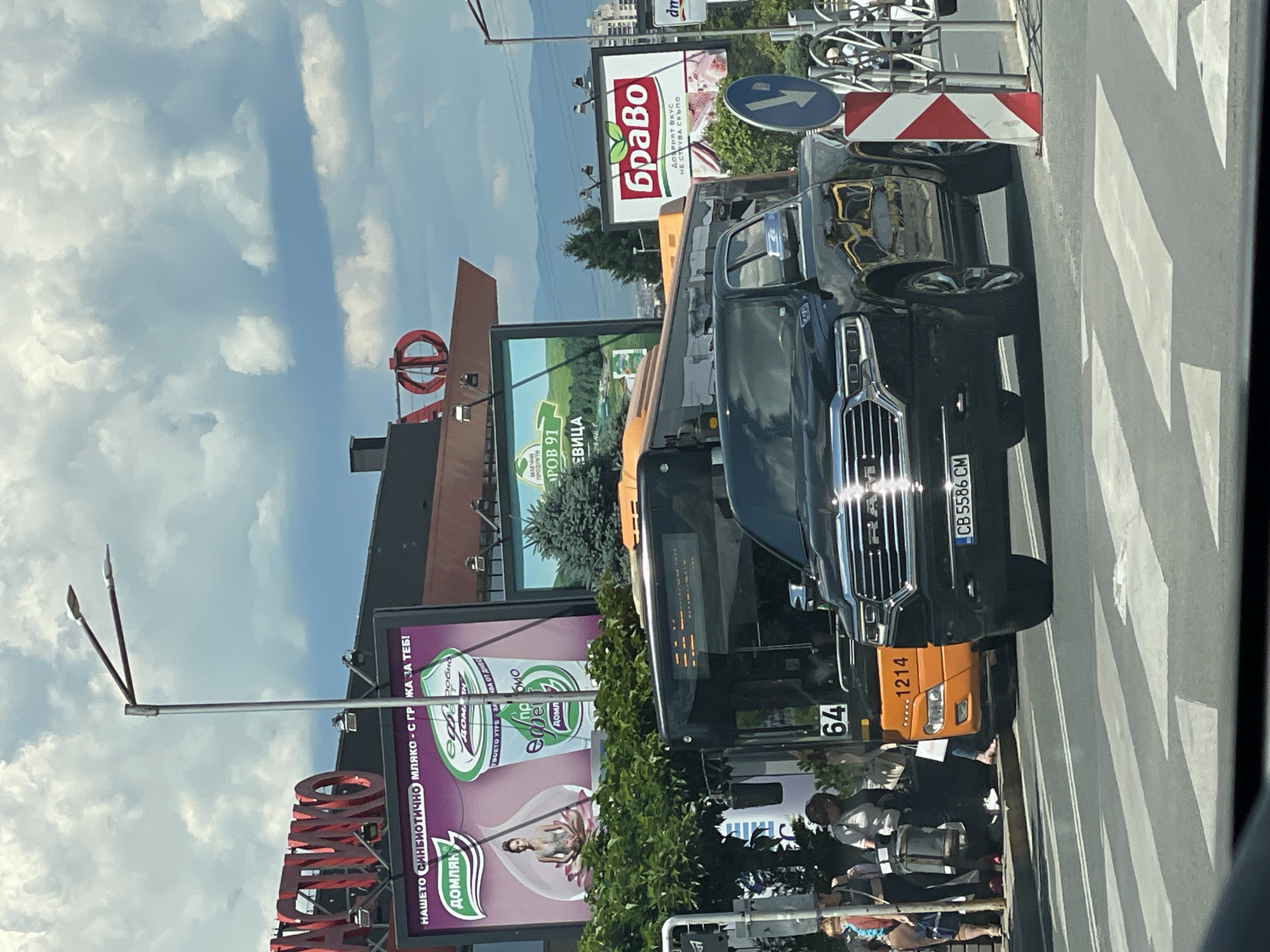 In this medium-shot, eye-level photograph, a black pickup truck is parked at a crosswalk in Sofia, Bulgaria. The truck is positioned in the foreground, facing right. Behind the truck, an orange and black bus is also stopped. Pedestrians are visible on the sidewalk to the left of the bus. Above and behind the vehicles, billboards and signage are displayed, including advertisements for "Domlyak" probiotic yogurt and "Bravo" meat products. The sky is cloudy, with a mix of blue sky and white clouds. Streetlights and buildings are visible in the background, indicating an urban environment. The overall scene depicts a moment at a busy city intersection on a bright, overcast day.