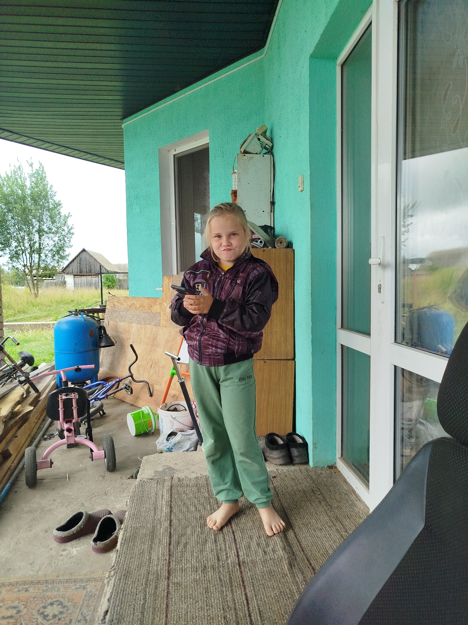 A young girl with blonde hair and a purple jacket stands on a porch in the village of Kryvaselski Sel'ski Sovet, Belarus. She is holding a black cellphone in her hands. She is wearing green sweatpants and is barefoot. In the background, there is a blue tank, bicycles, and a wooden house. The porch is covered by a green roof and has a turquoise wall.