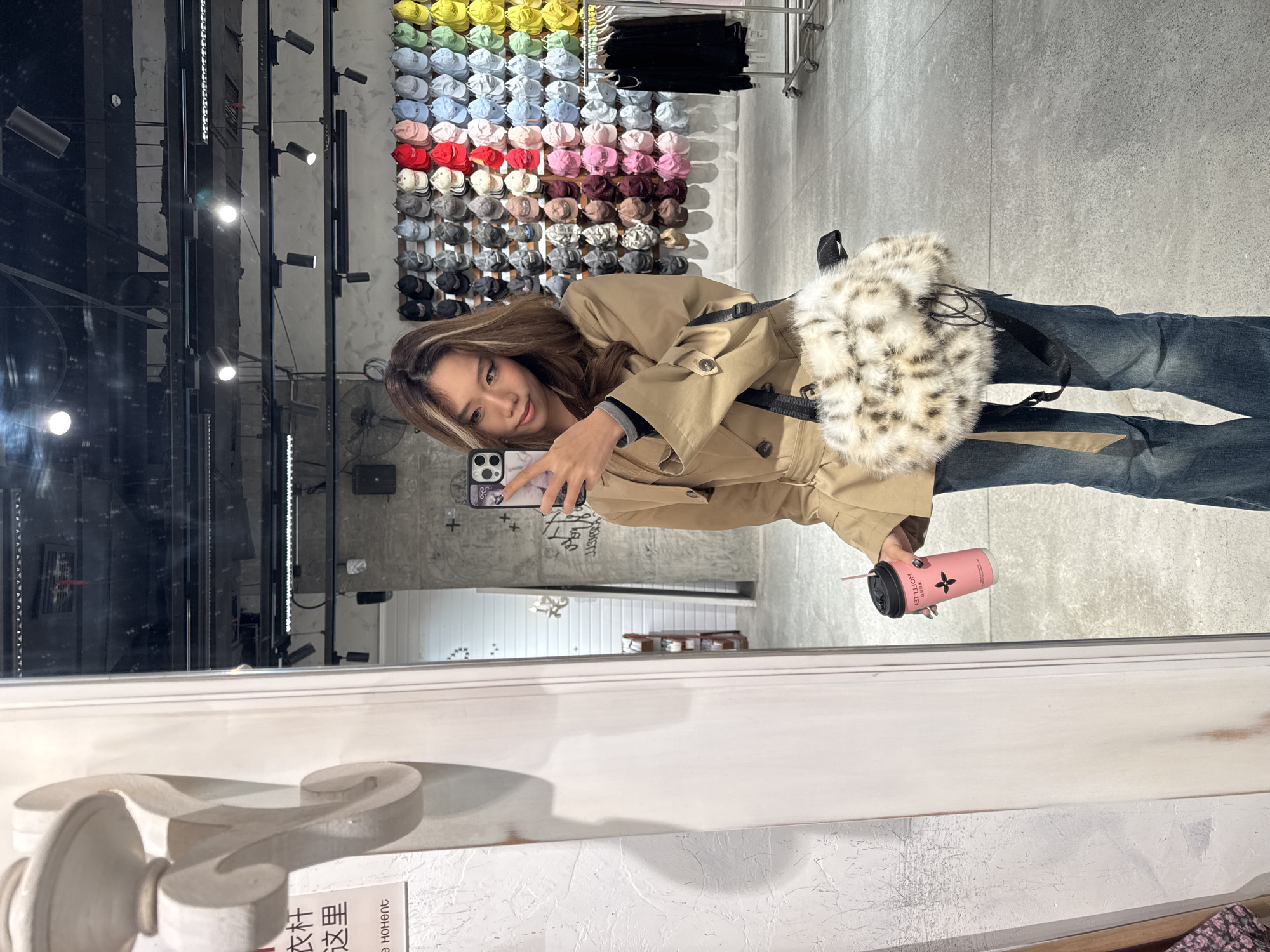 A person is reflected in a mirror holding a phone taking a selfie while holding a pink cup. The background features a wall of hats in various colors and a clothing rack with black garments. The location appears to be a clothing store or retail setting. Text in Chinese is visible at the bottom of the mirror frame. The person is wearing a tan trench coat and jeans. The cup has a straw sticking out of it. The image appears to be taken in the Jianlan Road District, China.