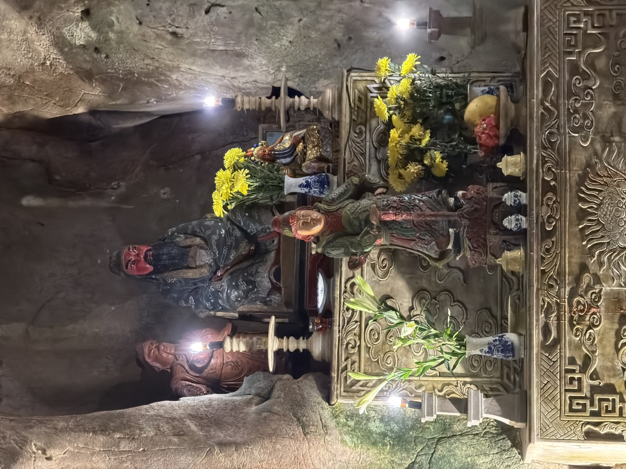 The image depicts an altar inside a cave in Đà Nẵng, Vietnam. Two statues stand at the top of the altar. One statue has a red face and a long black beard, while the other has a light orange face and is dressed in green. Yellow and white flowers in blue and white vases decorate the altar. A piece of fruit is on the altar as well. The wall behind the altar has carved patterns of clouds, dragons, and geometric shapes.