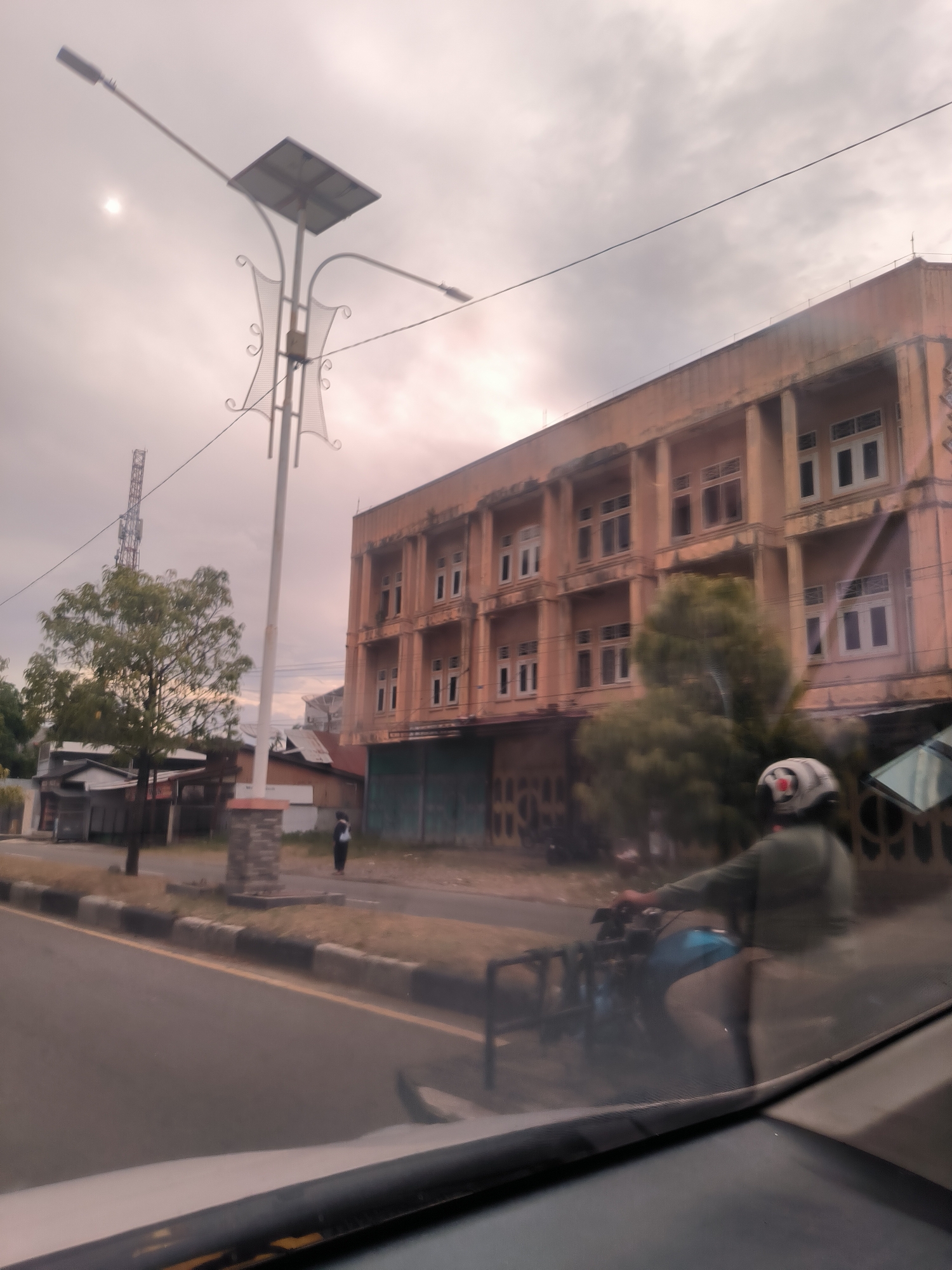 This image captures a daytime street scene in Sigli, Indonesia, under an overcast sky, with the sun faintly visible through the clouds in the upper left. The perspective is from inside a vehicle, with a portion of the dashboard and windshield visible at the bottom.
A prominent feature is a tall, decorative street light with a solar panel mounted on top, suggesting it is solar-powered. The light fixture itself is ornate, featuring intricate metalwork with scroll patterns.
To the right, a multi-story building dominates the background. It has a faded yellowish-orange or light brown facade, characterized by numerous white-framed windows and visible support columns. The ground floor features closed shutters, indicating it might be a commercial establishment.
On the left side of the road, a green tree partially obscures several smaller, simpler structures. A tall cellular tower is visible in the distant background.
The paved road features a yellow center line and a black-and-white painted curb. A person on a motorcycle is in motion in the foreground, wearing a white helmet and a green jacket, partially obscured by the vehicle's window and motion blur. Another person, dressed in dark attire and carrying a bag, stands on the dirt shoulder near the base of the street light, facing away from the camera.
The scene depicts a typical urban environment with a mix of modern infrastructure (solar street light, cell tower) and older architecture. No discernible text or signs are clearly visible.