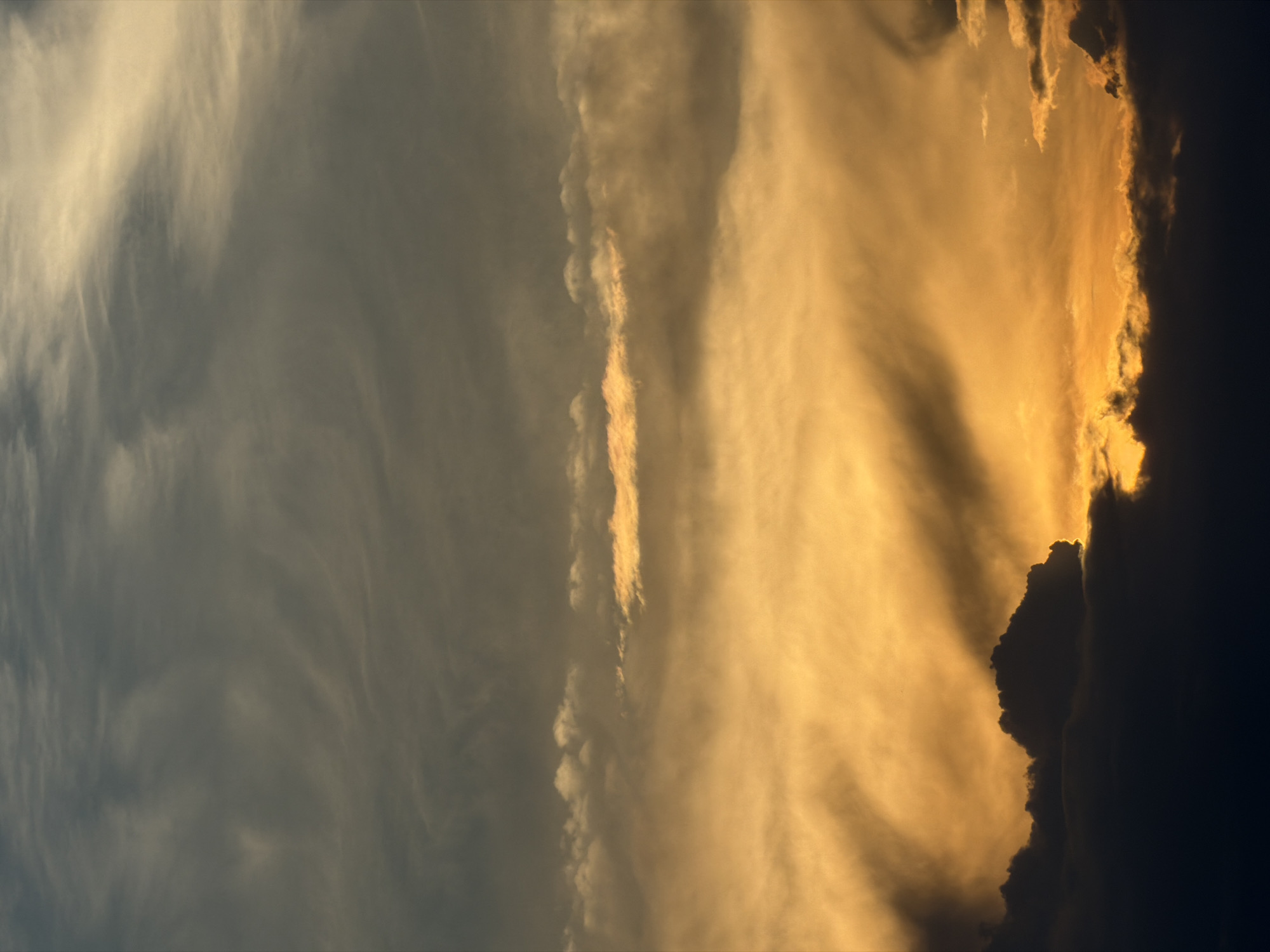 The image depicts a sky at dusk or dawn, with dramatic cloud formations. The left side of the image is dominated by bluish-grey clouds, while the right side is illuminated with a golden or amber hue, indicating the presence of the sun. There are dark, towering cloud formations on the right edge, creating a silhouette against the bright sky. There is no clear indication of the specific location or city, or of the presence of people, subjects, or objects.