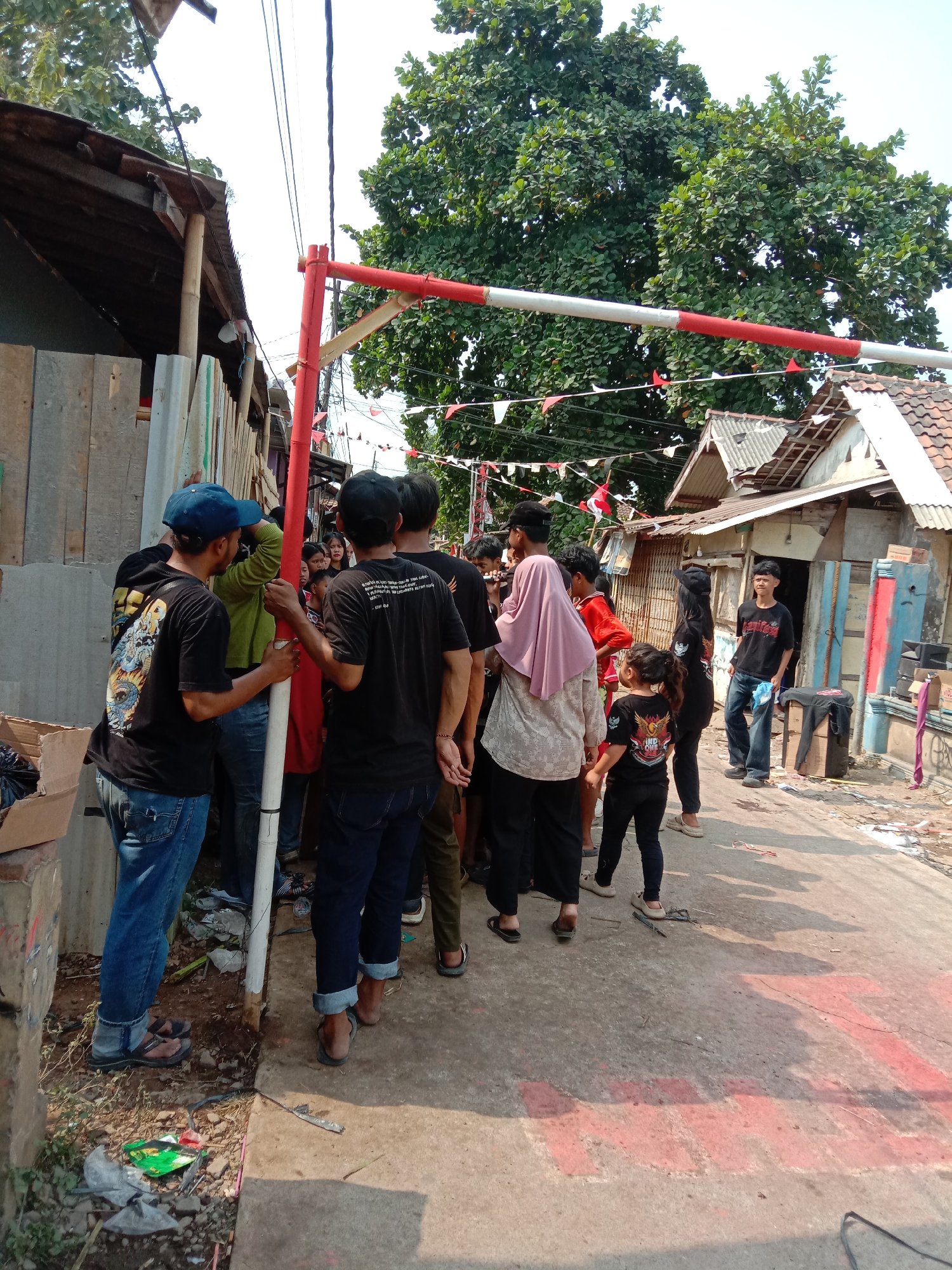 The image depicts an outdoor scene, likely in Karawang, Indonesia, characterized by a pathway lined with rudimentary structures. A group of individuals, primarily young males dressed in casual attire such as t-shirts and jeans, are gathered. Some are wearing black t-shirts with graphic designs. One individual wears a blue baseball cap, while another sports a pink hijab. A young girl in a black t-shirt and black pants is also present, as is a person wearing a black headscarf.
Dominating the foreground is a red and white striped barrier, resembling a gate, that appears to be temporarily erected. It is supported by red poles. Decorative red and white flags are strung across the scene, suggesting a festive occasion or a local event. The pathway itself has red markings painted on it, possibly indicating a boundary or a designated area.
The environment consists of a rough concrete pathway with dirt and debris along its edges. On one side, there are wooden structures with corrugated metal roofs, indicating informal housing or shops. On the other side, more buildings with tiled roofs are visible, some appearing weathered. Lush green trees with dense foliage provide a backdrop, suggesting a tropical or subtropical climate. The presence of electrical wires overhead further signifies an inhabited area. The overall impression is one of a vibrant, informal community space possibly decorated for a local celebration.