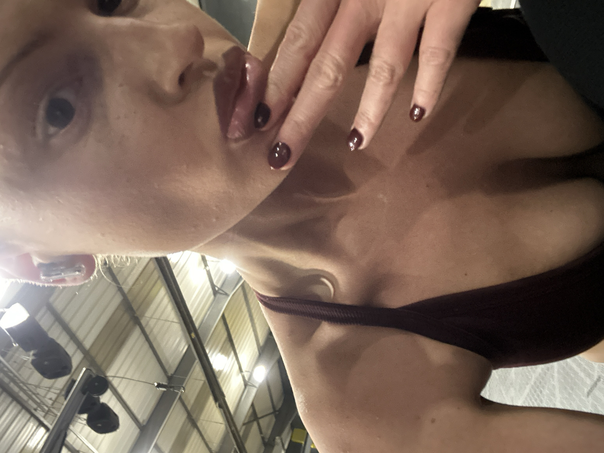A selfie-style photograph, likely taken indoors given the industrial-style ceiling structure in the background. The subject is a person with fair skin and dark nail polish, visible on the fingers that are near their face. The person's lips are glossy. The subject appears to be wearing a dark sports bra or top. The location in the city of Tonbridge and Malling, United Kingdom is unknown.