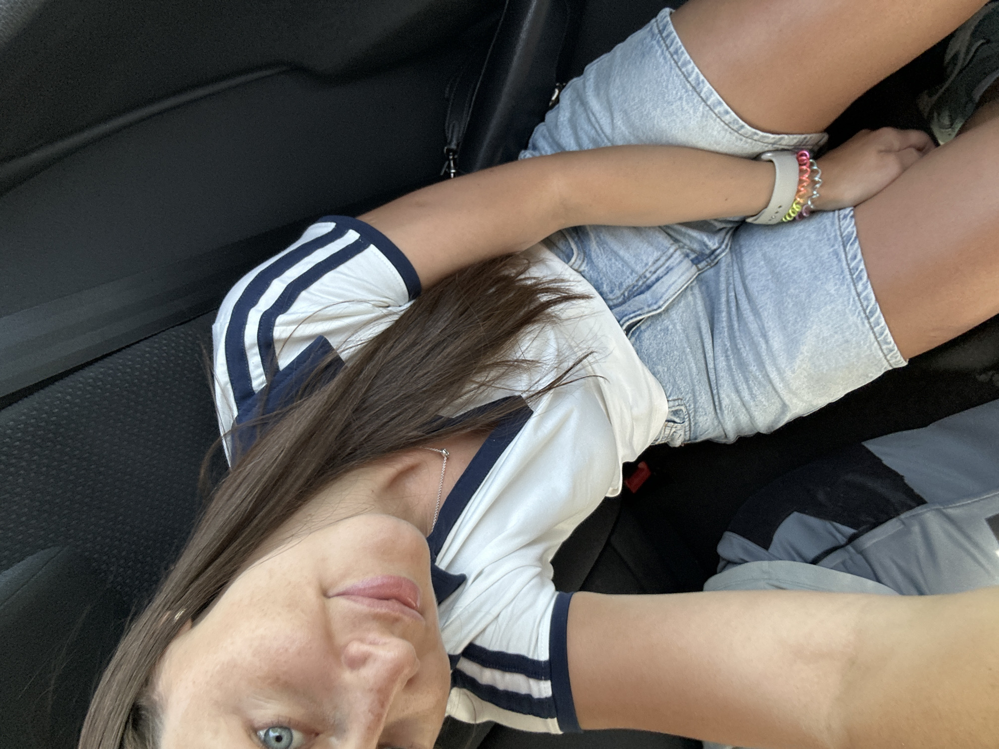 A person is seated in what appears to be the driver's seat of a car, wearing a white t-shirt with dark blue stripes on the sleeves and collar, and denim shorts. The person's left arm rests across their legs, with a multi-colored bracelet visible on their wrist. The car's interior is dark, suggesting a recent model. The image provides no information to suggest that this scene is in Walsrode, Germany. Therefore, I cannot verify this location.