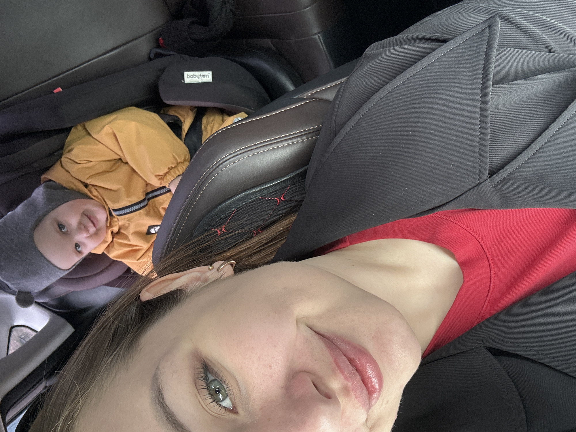 The image shows a person and a child in a car, possibly in Chaikovsky, Russia. The adult, wearing a red shirt and a black blazer, is positioned closer to the camera, while the child, wearing a grey hat and yellow jacket, is in a car seat in the background. A baby carrier, labeled "babyton," is also visible near the child. The car's interior, including the brown leather seat, is captured.