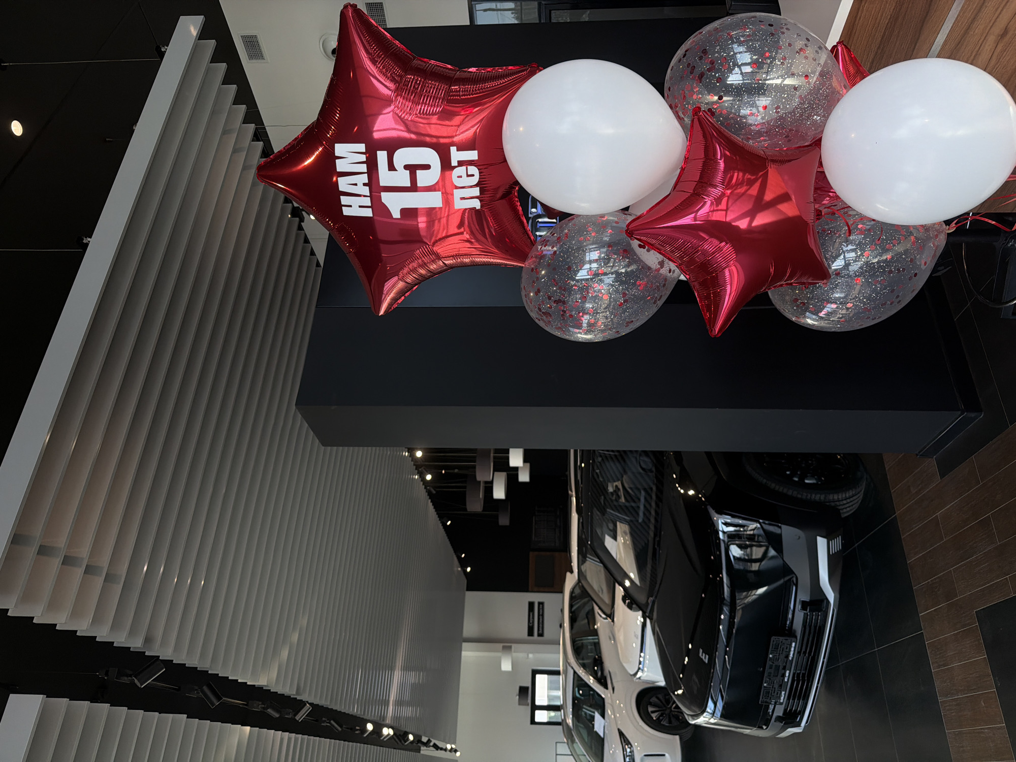 The image depicts a car dealership, possibly celebrating its 15th anniversary. The location is likely in Izhevsk, Russia, based on the Cyrillic lettering "НАМ 15 лет" (meaning "We are 15 years old").
The scene includes:
* A cluster of balloons: two red star-shaped balloons with the celebratory inscription, two clear balloons filled with confetti, and two white balloons.
* Two vehicles parked closely together, a white SUV and a black SUV.
* A modern interior with a dark ceiling and light-colored, slatted walls.
* Track lighting fixed on the ceiling.