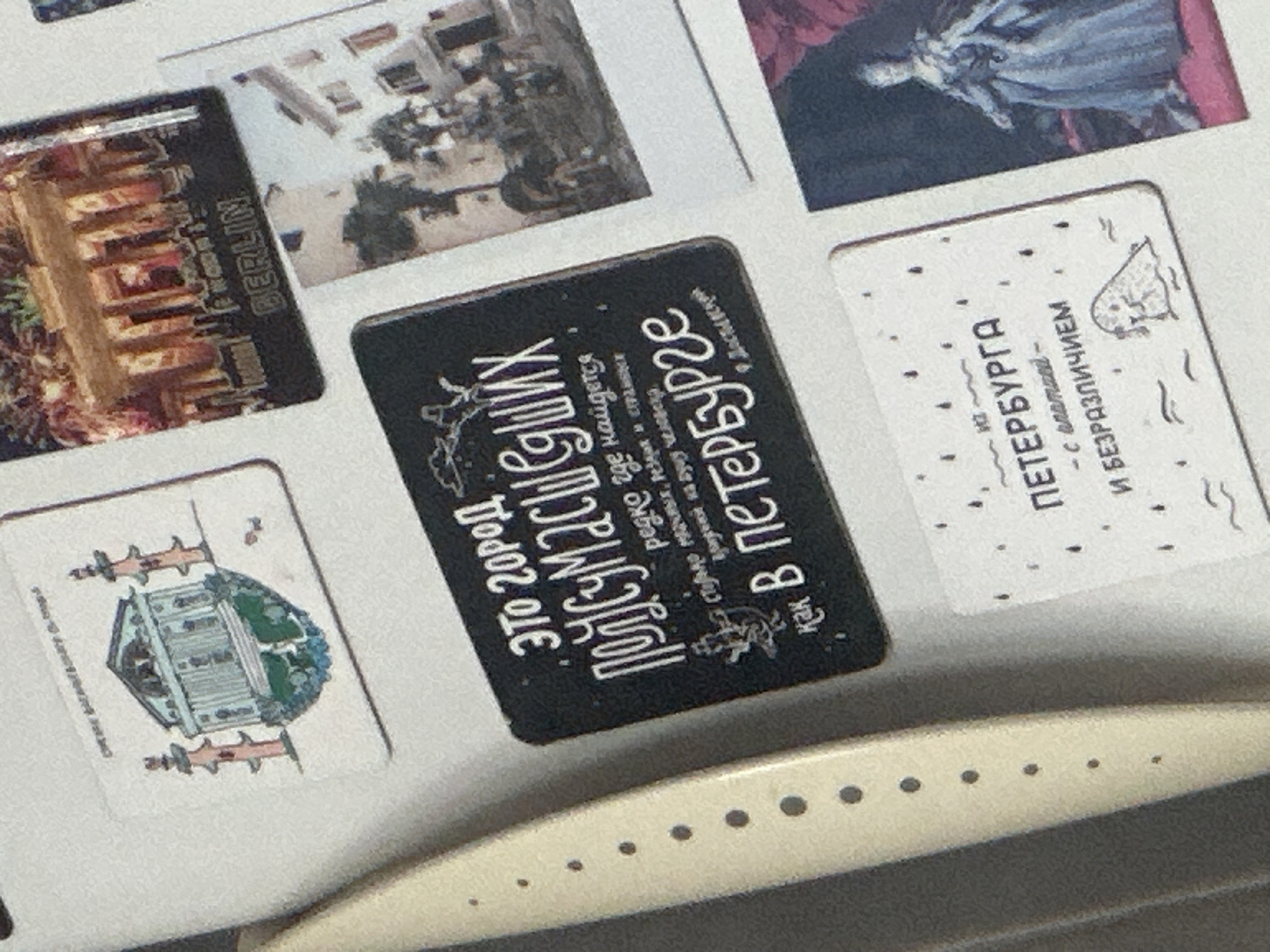 This media file showcases a collection of postcards or magnets arranged on a white surface, possibly a refrigerator or a bulletin board. The items feature various urban landscapes and text, primarily in Russian.
One prominent item is a black rectangular magnet with white text in a stylized font. The text reads "Это город Полусумасшедших редко где найдется столько мрачных, резких и странных предний на душу человека. как В Петербурге." This translates to "This is a city of the half-crazy, rarely will you find so many gloomy, sharp, and strange whims on a person's soul. How in Petersburg." The magnet also depicts a silhouette of a person or creature.
Another item is a white rectangular postcard with a drawing of a building that appears to be an institutional structure with columns and a dome, surrounded by trees. Text is present in both Russian and English. The Russian text reads "ПЕТЕРБУРГА - с опатией - И БЕЗРАЗЛИЧИЕМ", which translates to "OF PETERSBURG - with apathy - AND INDIFFERENCE." The English text is partially obscured but appears to be the name of the building.
A third item, a postcard or magnet, features a vertical inscription "BERLIN" next to an image of a large building with intricate architecture.
The overall impression is one of travel souvenirs or personal mementos, possibly related to trips to Berlin and St. Petersburg. The lighting suggests an indoor setting, with no specific time of day or weather conditions evident. The arrangement is somewhat haphazard, indicating a casual display.