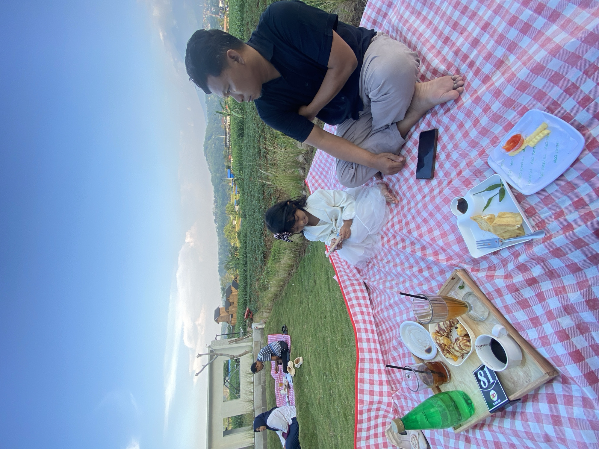 This is a photograph taken at a picnic area in the city of Kesiman, Indonesia, with a view of rolling green hills and a blue sky with scattered clouds. In the foreground, a man and a young girl are seated on a red and white checkered picnic blanket. The man is wearing a black polo shirt and light-colored pants, with his left arm crossed over his chest. The girl is wearing a white dress and appears to be looking down at something in her hands.
On the blanket in front of them, there is a wooden tray with various food and drinks, including a green bottle, two glasses of dark liquid, a white cup of coffee, and a plate of what appears to be fried food. There is also a smartphone lying on the blanket near the man's feet.
In the mid-ground, further back on the grass, another picnic scene is visible. A woman in a blue hijab and a white shirt, and a man in a striped shirt are seated on a picnic blanket.
The background shows lush green fields, some structures that appear to be buildings, and trees, all set against a backdrop of distant hills and the sky. The overall scene depicts a relaxed outdoor gathering with food and drinks enjoyed in a natural setting.