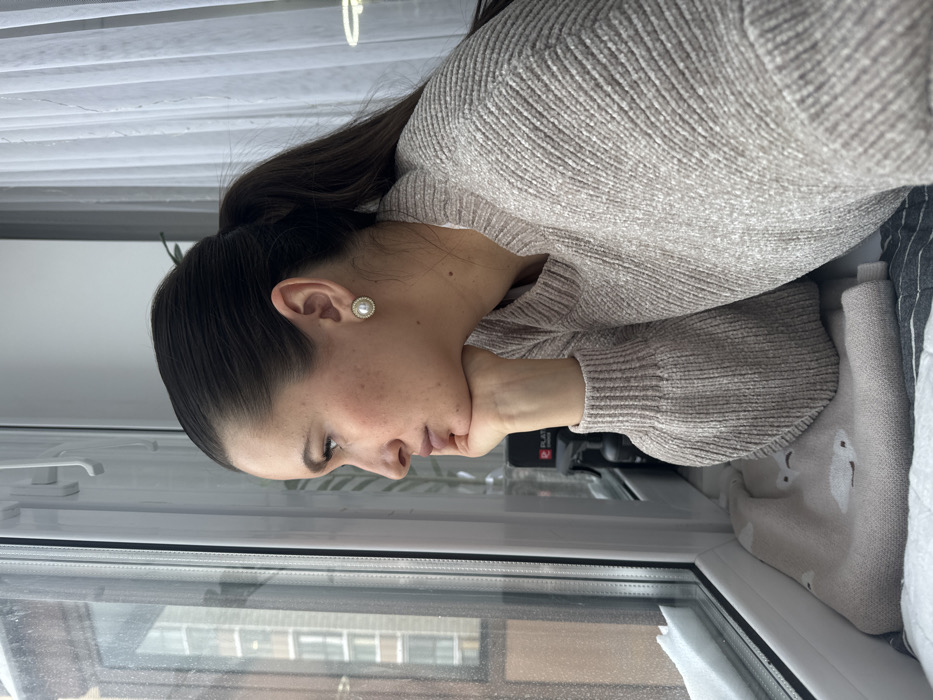 A female subject is depicted in profile, facing left, looking out a white PVC window in Odintsovo, Russia. She has dark hair tied back in a ponytail and is wearing a light beige textured knit sweater and round pearl earrings with a gold-colored rim. Her right hand supports her chin. Outside the window, a multi-story building is visible, and snow is present on the exterior windowsill. Inside, on the windowsill, a light beige hot water bottle cover with white cloud patterns rests on a dark grey and white striped fabric. A dark object with a red "PLAT" logo is visible behind the subject's hand on the sill, along with white sheer curtains and a hint of a green plant in the background. Natural daylight illuminates the scene.