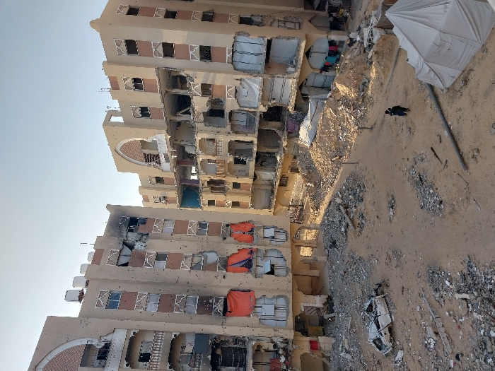 This image depicts a severely damaged urban landscape in Khan Yunis, Palestinian Territory, under a clear, bright sky. Two multi-story, light-colored buildings with arched architectural elements are prominent. The building on the left exhibits numerous impact holes on its facade, shattered windows, and significant black scorching around its upper floors. Orange tarpaulins cover several lower-level window openings. The building on the right shows extensive structural compromise, with large sections of its exterior walls missing, revealing multiple damaged internal floors. White tarpaulins or fabric partially cover some openings on this structure, and laundry is visible hanging near its ground level. The unpaved ground between the buildings is covered with sand, dirt, and significant amounts of debris, including broken concrete and scattered rubble. A severely damaged white vehicle chassis, stripped of its wheels, lies on its side in the lower-left portion of the frame. A single individual, dressed in dark clothing, walks across the debris-strewn ground towards the right-center of the image. A large, light-colored tent is pitched on the sandy ground in the lower-right foreground. A substantial pile of rubble and dirt is present in the middle-right area.