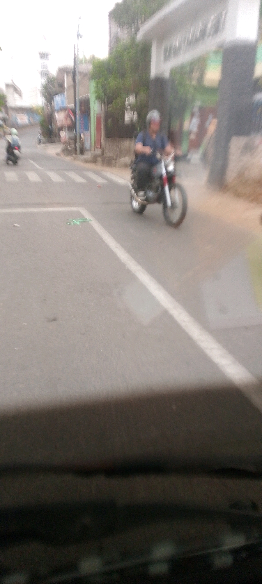The image depicts a street scene in Bandung City, Indonesia, as evidenced by the architecture and the presence of a motorcycle with a rider. A man wearing a blue shirt and a helmet is riding a motorcycle on a paved road. The motorcycle is partially visible, showing its wheels and exhaust pipe. A pedestrian crossing is marked on the road. In the background, there are buildings with some greenery and trees. The sky is overcast. The image is blurry, suggesting motion or a low-quality camera.