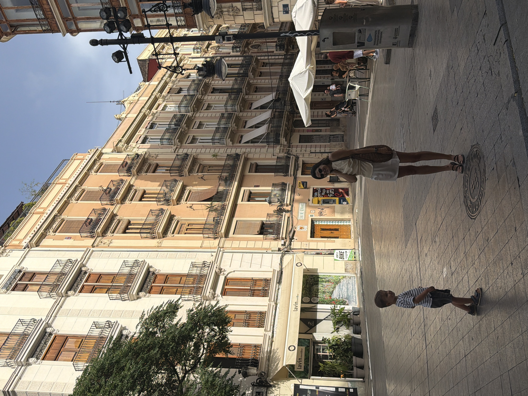 A full shot shows a woman and a child standing on a street in front of buildings. The woman is in the foreground, wearing a white skirt and a light-colored top. She is smiling and touching her face, looking towards the camera. A woven bag hangs from her shoulder. The child, a young boy, is to her left and slightly further back, wearing a striped t-shirt and shorts. He is looking to his right. Behind them, various buildings line the street. The buildings are multi-story with balconies and ornate architectural details. In the background, there are people sitting at tables outside a cafe with white umbrellas. The street is paved with cobblestones. A lamppost with multiple lights is visible to the right. The scene is lit by sunlight, casting shadows on the ground.