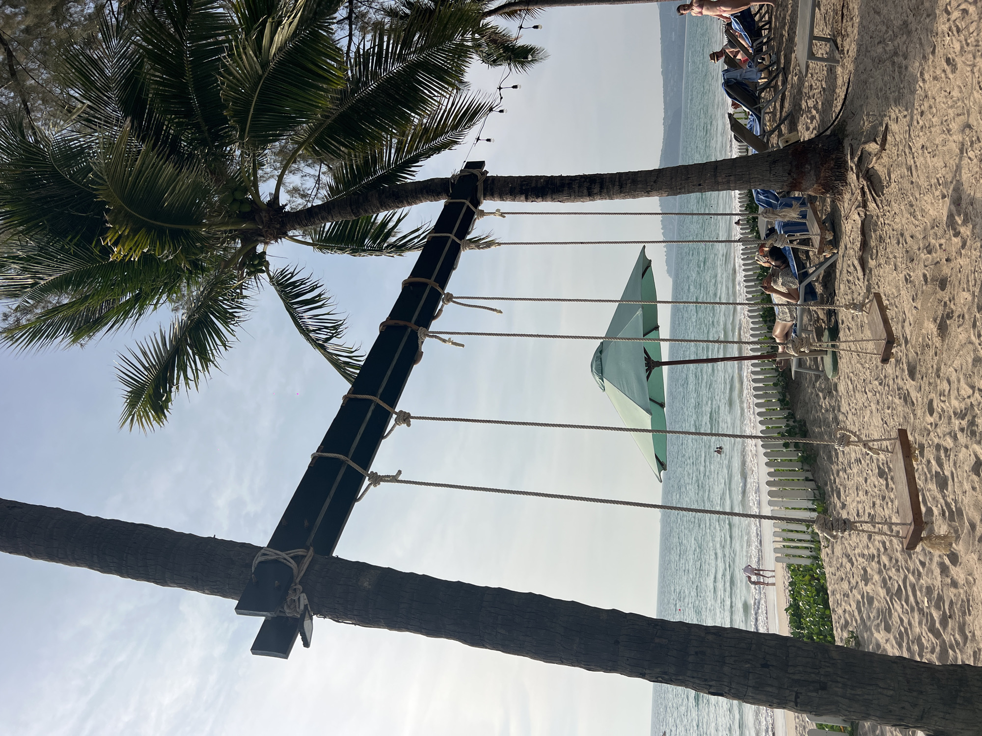 The image depicts a beach scene in Choeng Thale, Thailand. A swing is suspended between two palm trees, with ropes securing it to a wooden beam. An open, green umbrella is positioned near the swing. In the background, the ocean is visible, with people lounging on beach chairs and enjoying the water. A white picket fence runs along part of the beach, and some vegetation is visible. The sky is light, suggesting a daytime scene.