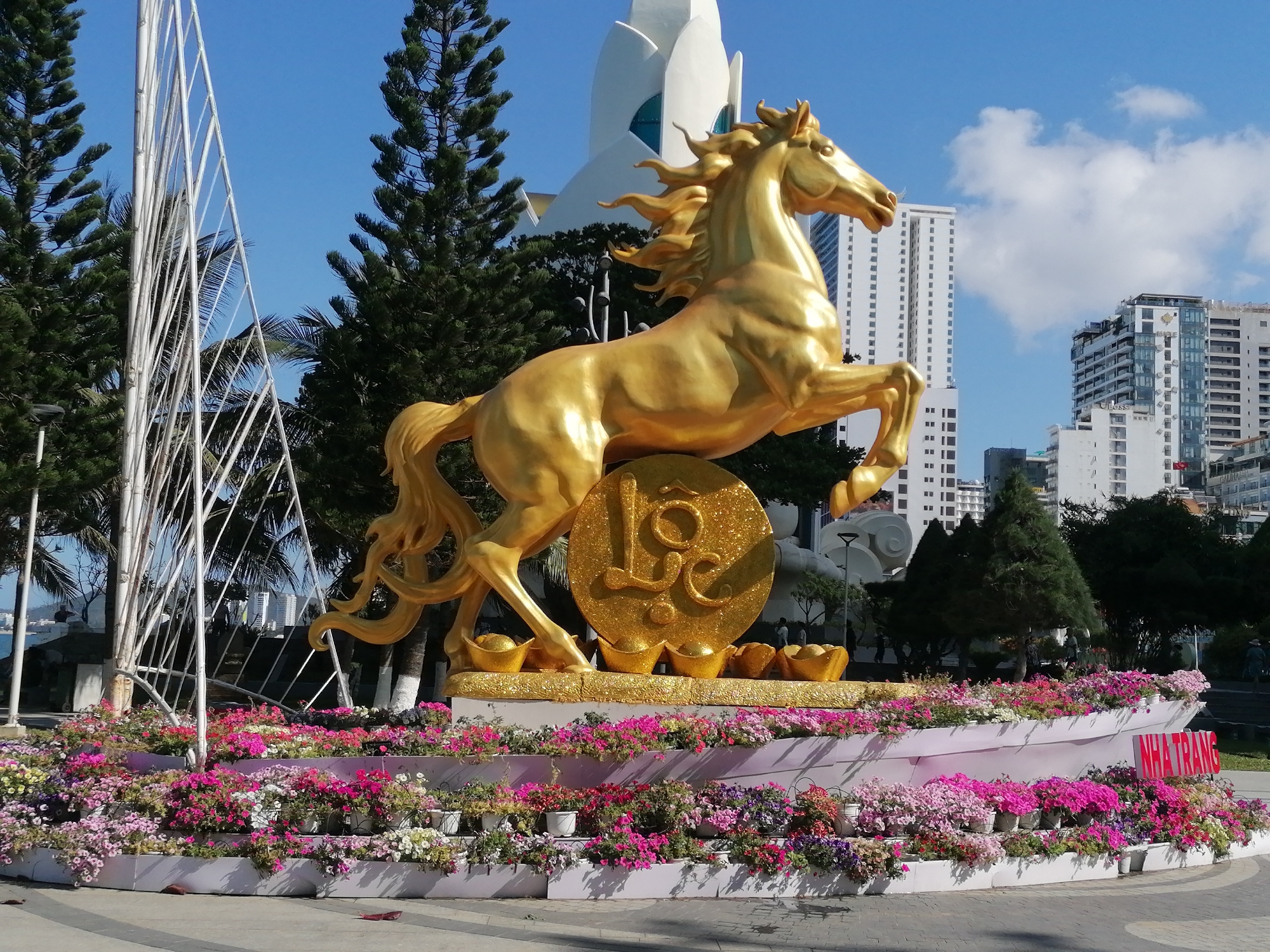 The image shows a golden horse statue in a public square, likely in Nha Trang Ward, Vietnam, based on the inscription "NHA TRANG" on the flower display. The statue is positioned on a tiered platform decorated with flowers in various shades of pink, white, and purple. The horse is rearing on its hind legs, with a golden disc bearing the inscription "LOC" behind it. In front of the horse are golden, boat-shaped figures.
Behind the statue are several evergreen trees and a modern, white architectural structure. Further in the background, are high-rise buildings under a blue sky with scattered clouds. To the left of the statue is a white metal framework structure. The ground around the statue and flower display is paved.