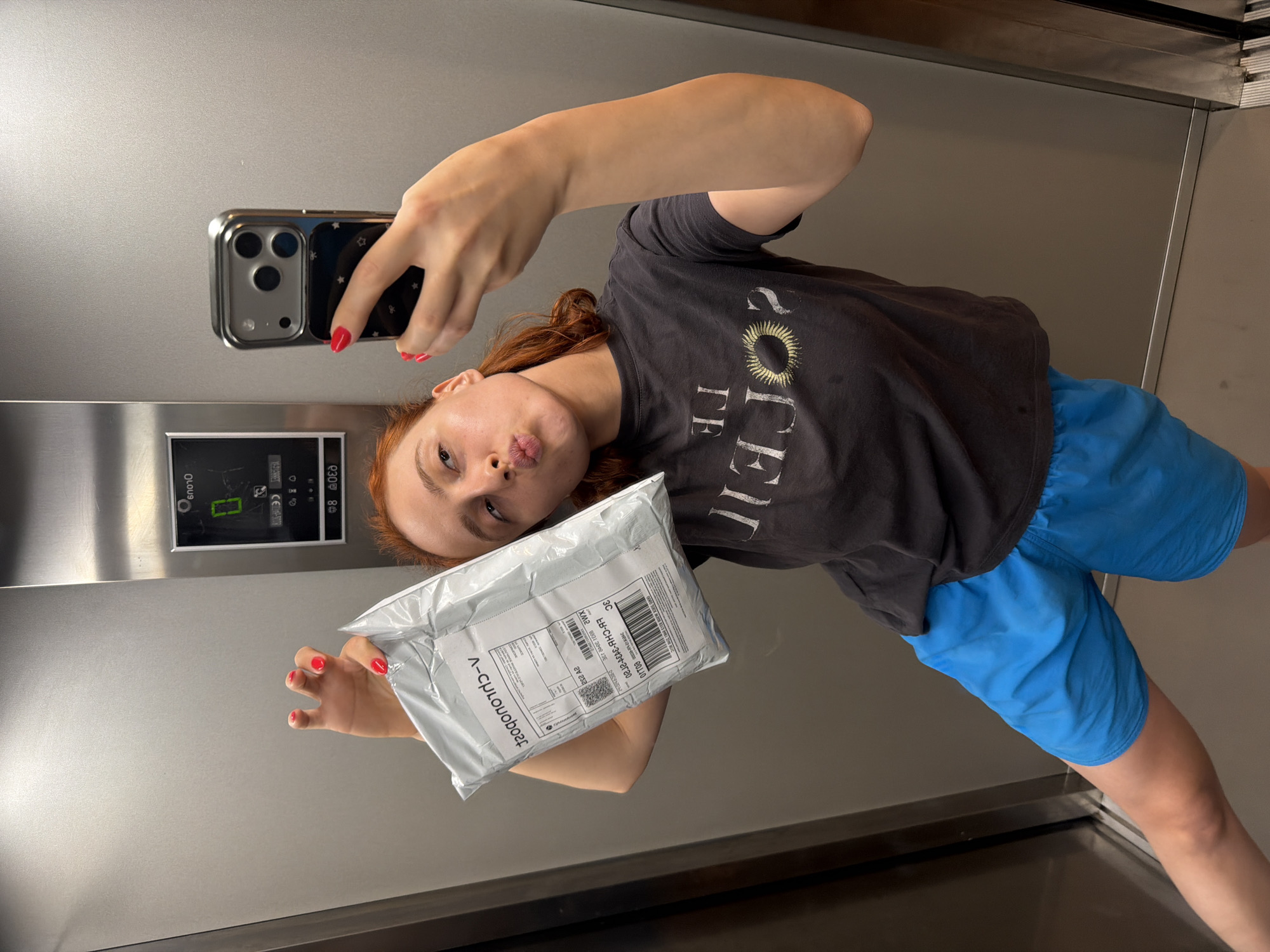 In an elevator, a young woman with auburn hair, wearing a dark grey t-shirt with "THE SOLE" printed on it and bright blue shorts, poses for a selfie. She holds a padded envelope package in her left hand, which is extended upwards and angled towards the camera. Her right hand holds a smartphone, taking the picture. The woman is puckering her lips, and her nails are painted a vibrant red. The elevator walls are metallic and grey, with an elevator control panel visible in the background. The scene takes place in an indoor environment, possibly a residential building or hotel elevator. The specific location of Strasbourg, France, is not indicated by any visual cues in the image.