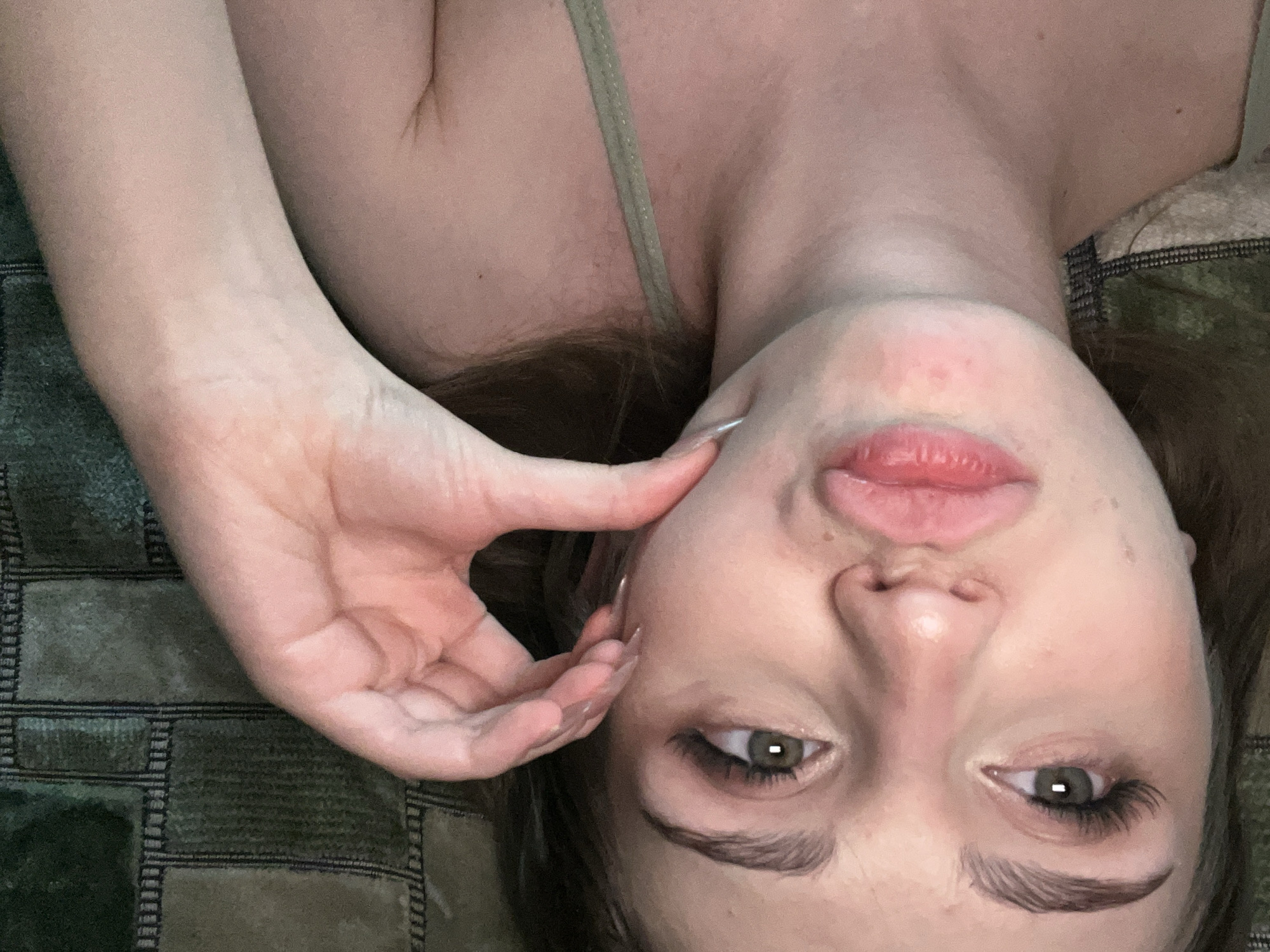 A close-up, upside-down portrait of a person lying on their side. Their head is tilted and their face is turned towards the viewer, with one hand touching their cheek. The person has fair skin, green eyes, and full lips that are slightly parted. Their eyelashes are long and dark, and their eyebrows are well-defined. They are wearing a light-colored strap, possibly from a camisole or bra. The background is a dark, textured surface with a geometric pattern in shades of green and brown. The lighting appears to be soft, with no harsh shadows. The image has a candid and intimate feel, suggesting a relaxed or playful moment. There is no visible text in the image. The location is Ashkadar, Russia.