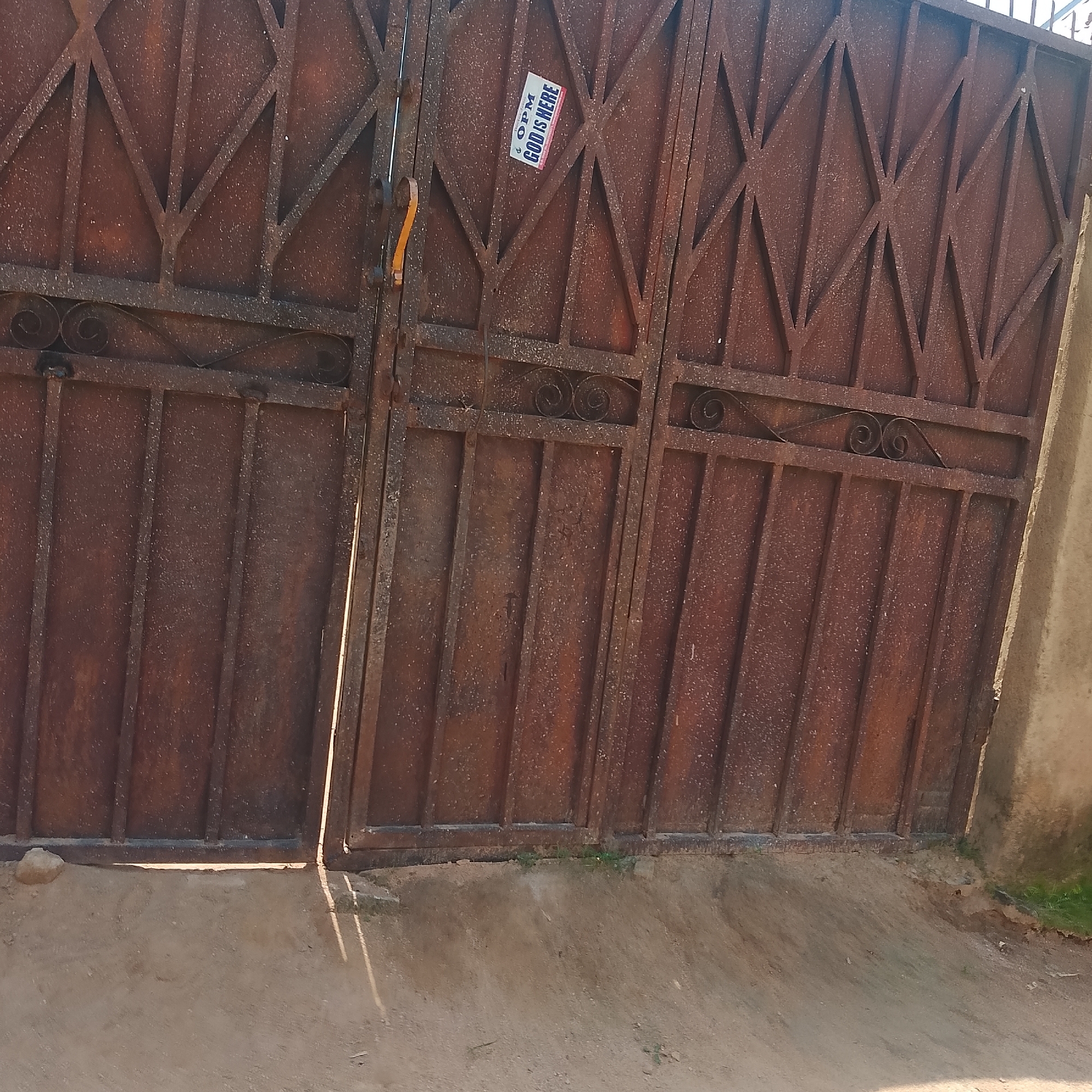 This is a close-up, slightly angled shot of a weathered, rusty metal gate. The gate is composed of vertical bars and a decorative diamond pattern at the top. A small, rectangular sticker is affixed to the right panel of the gate, slightly askew. The sticker has text that reads "OPM GOD IS HERE" in white and red lettering on a blue background. The gate appears to be in an outdoor setting, with a sandy or dirt ground in the foreground. The sunlight casts shadows on the ground, suggesting it is daytime. The overall impression is one of an aged, functional object in a simple, possibly rural or semi-urban environment.