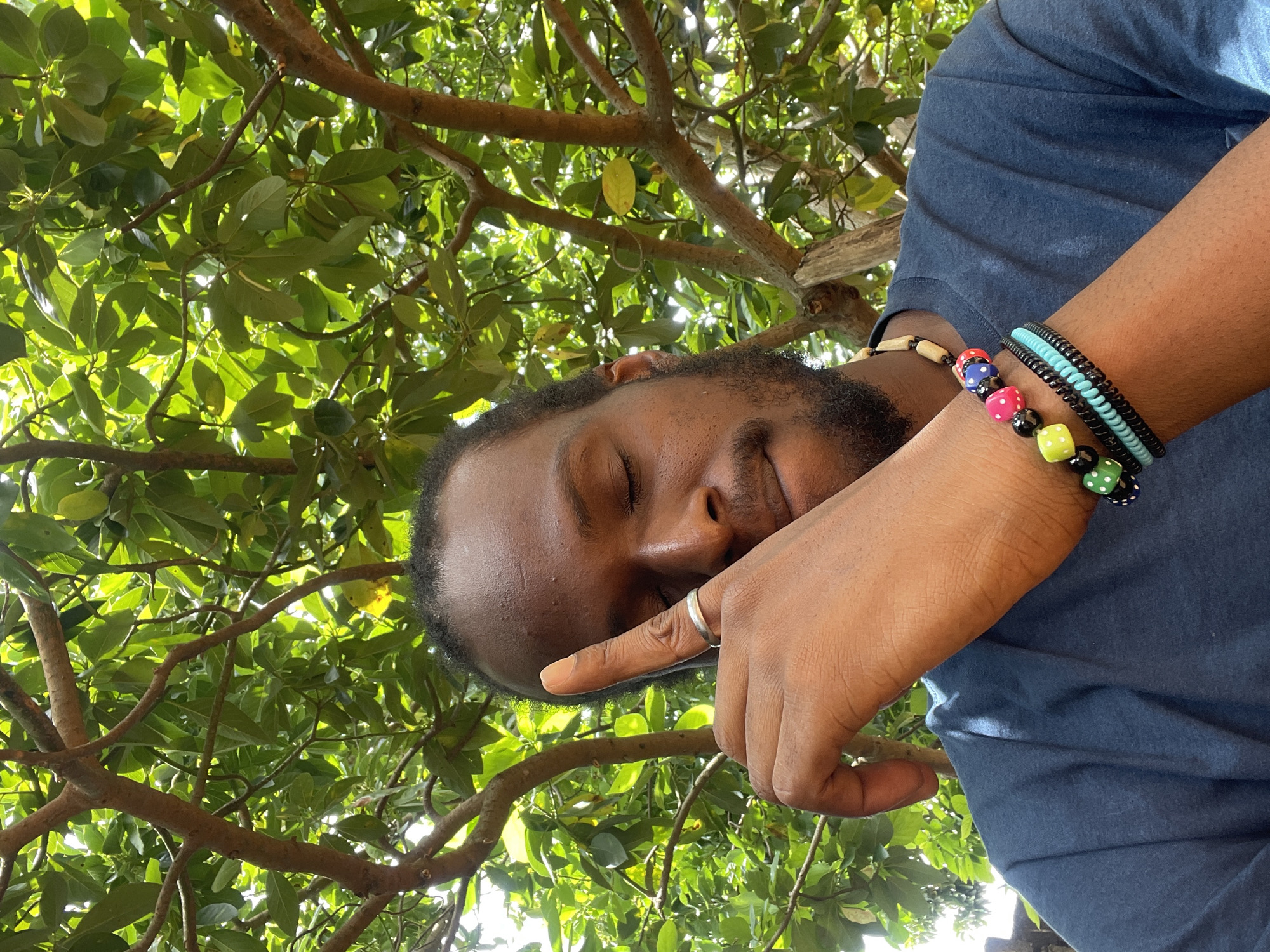 A man is photographed in Jos, Nigeria, possibly outdoors under a tree. He is wearing a blue shirt and has multiple bracelets on his right wrist, one made of dice beads. His eyes are closed, and his right hand is raised with his index finger near his eye. A silver ring is visible on his finger. The background consists of tree branches and green leaves, with sunlight filtering through.