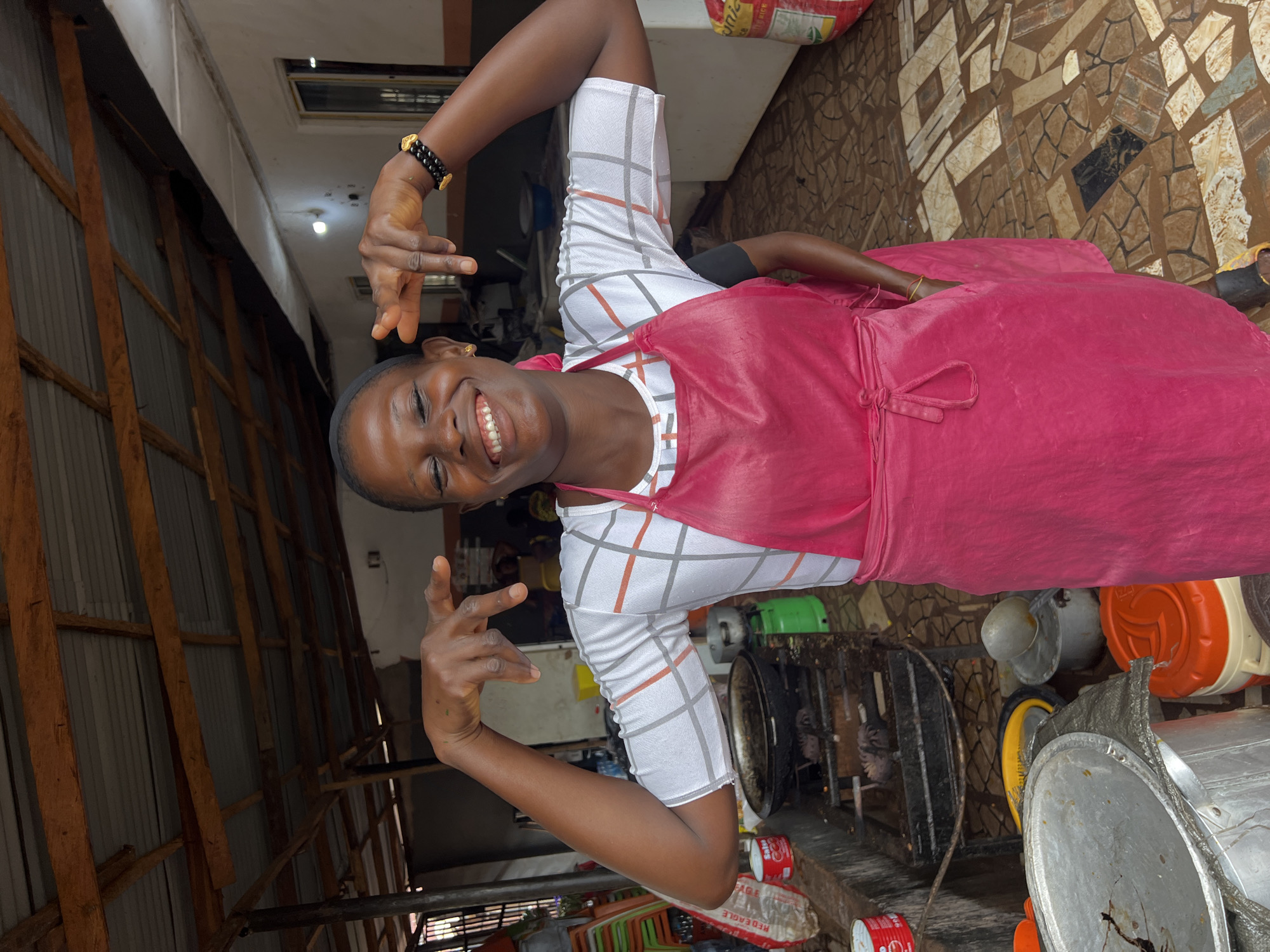 The image depicts a woman in Nkpor, Nigeria, wearing a white and grey checkered shirt and a bright pink apron. She is standing in what appears to be an open-air food stall or kitchen area. Her eyes are closed, and she is smiling broadly, with her hands raised in a playful gesture. She has a black headband on and a beaded bracelet on her right wrist. Behind her, a stove with cooking pots is visible, along with various containers and sacks, suggesting a busy food preparation environment. The ground is paved with stone tiles. The overall atmosphere appears lively and cheerful.