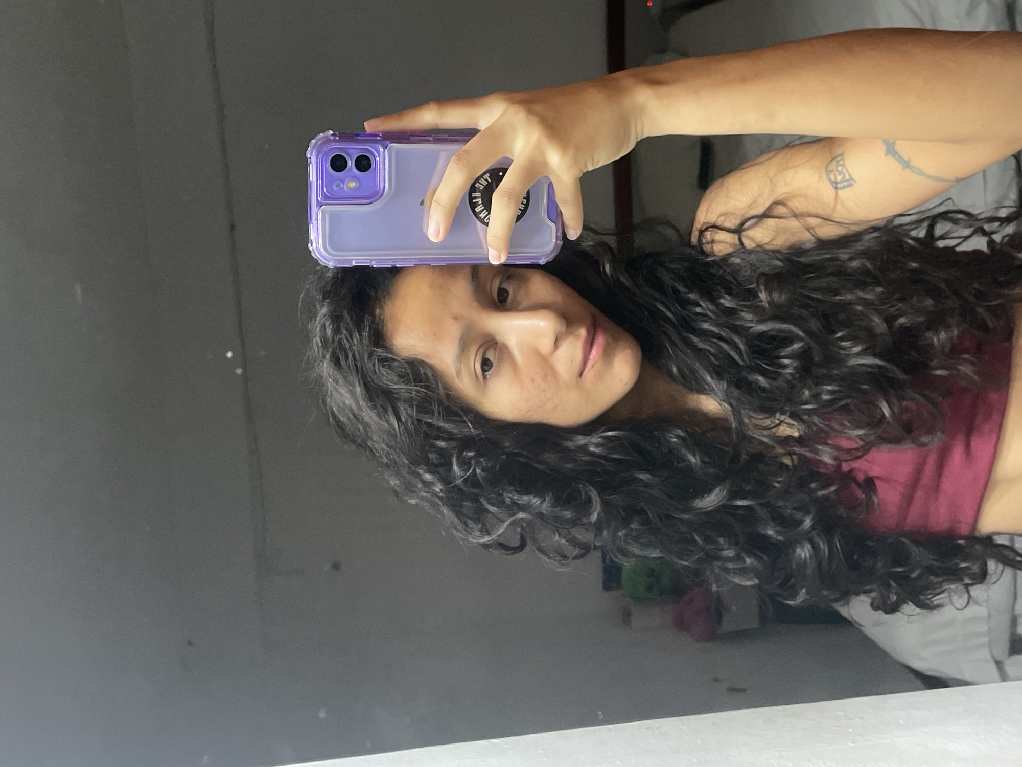 In Porlamar, Venezuela, a young woman with long, dark, curly hair is taking a selfie in a mirror. She is wearing a maroon crop top and has a purple phone case on her phone, which she is holding up to take the picture. The background is a dimly lit room with a white wall on the left and a bed with gray bedding in the background. There are some objects on a shelf in the background, including a green stuffed animal and a pink item. The woman has visible acne on her face and a tattoo on her left arm. The overall scene is casual and personal, suggesting a moment captured in a private space.