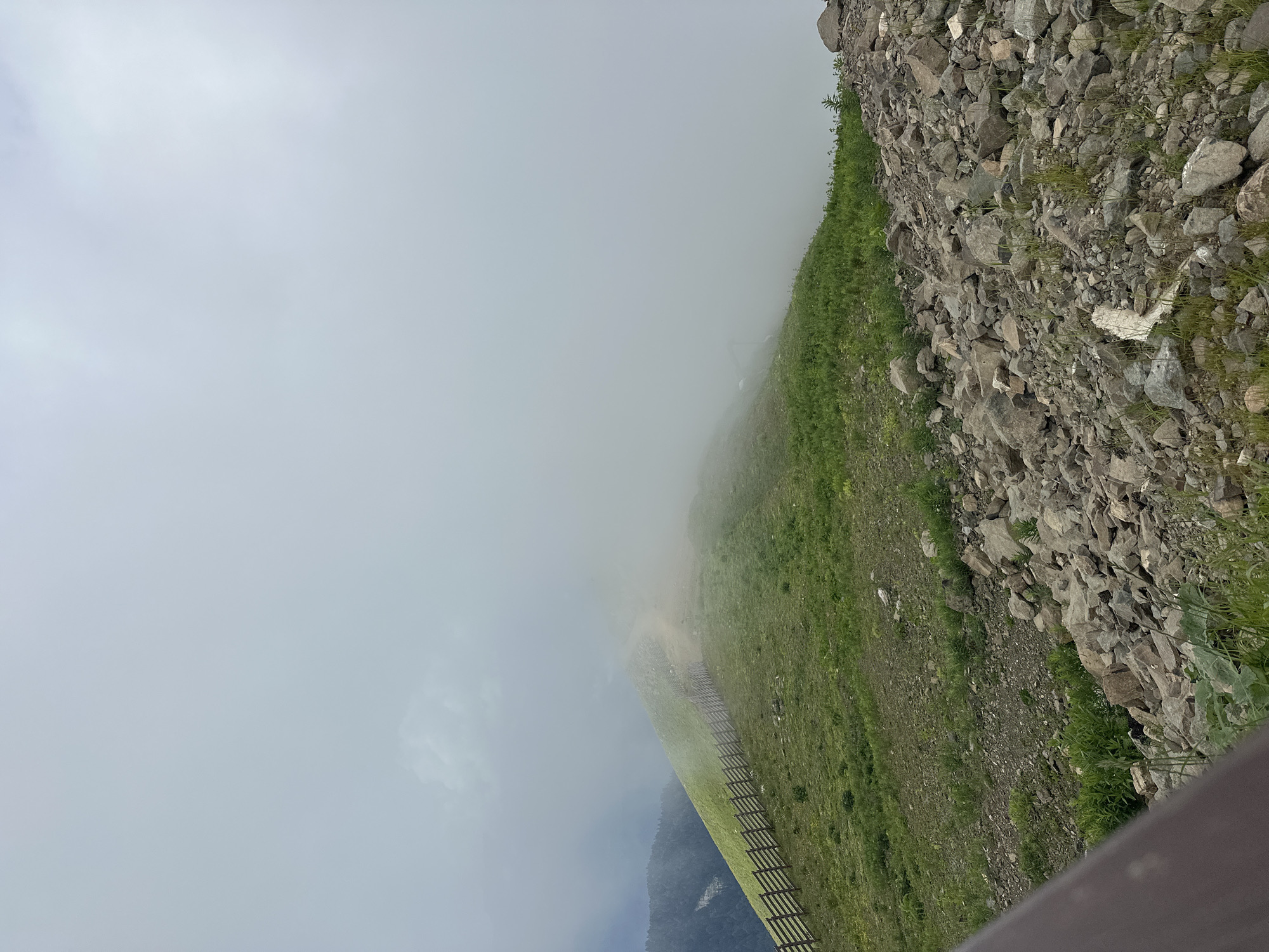 The image depicts a mountain ridge covered in green grass and rocks, shrouded in thick fog. A wooden fence runs along the ridge, marking a path that disappears into the mist. In the foreground, a pile of rocks is visible, along with sparse vegetation. The scene appears to be from a high-altitude location, possibly a hiking trail or a viewpoint in Estosadok, Russia, given the context of the surrounding mountains. The overall atmosphere is misty and overcast, suggesting a cool or damp day. There are no people or animals visible in the frame.