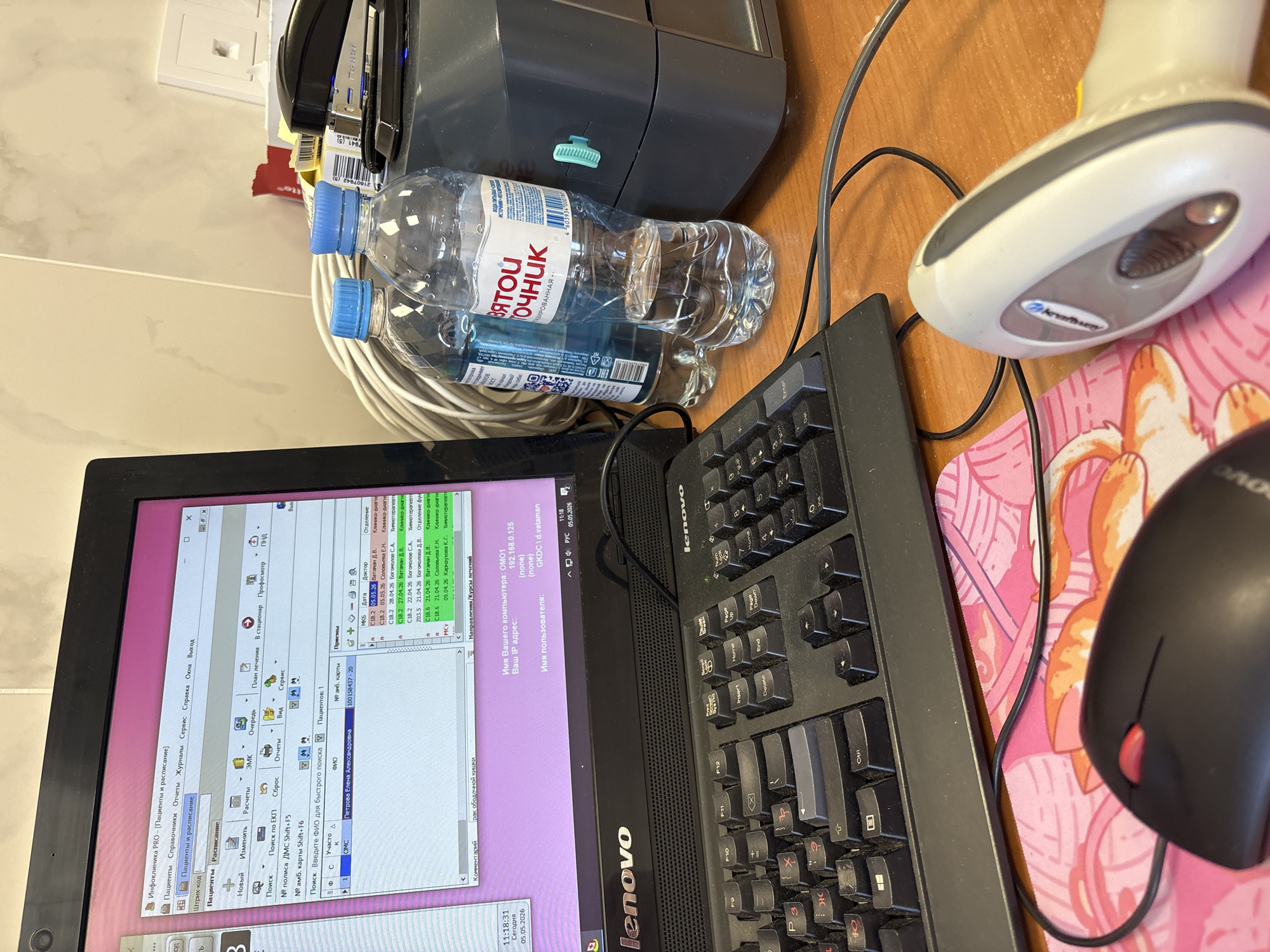 The image depicts a cluttered workspace with a laptop displaying Russian text, a barcode scanner, a computer mouse, a keyboard, two bottles of "Svyatoy Istochnik" (Holy Spring) mineral water, a printer, a stapler, and several cables. The laptop screen shows a program labeled "Infoclinica PRO" with patient records and scheduling information. The computer's IP address is 192.168.0.125 and the username is "GKDC d.vataman". The date displayed on the screen is May 5, 2026. The bottles of water are labeled in Russian with the name "Svyatoy Istochnik" (Holy Spring). The keyboard is a Lenovo brand, and the barcode scanner is a handheld device. A black computer mouse is positioned on a pink mousepad featuring a pattern with orange and white cat-like figures. The location is implied to be in Russia because of the Russian text and the Russian brand of water. There are no indications of Saint Petersburg in the image.