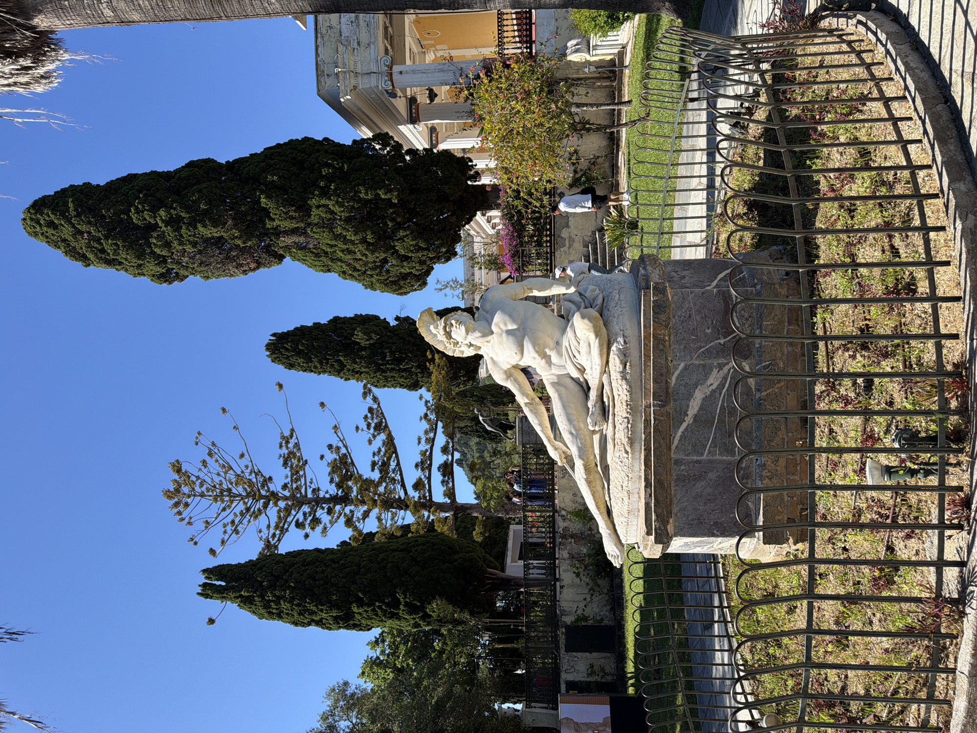 This vibrant outdoor scene depicts a classical garden or palace grounds under a clear blue sky, likely in Gastouri, Greece. In the foreground, a prominent white marble statue of a male figure is mounted on a stone pedestal. The figure is shown in a dynamic, reclining pose, possibly depicting a hero or mythological character.
A dark metal fence with decorative curved tops runs across the middle ground, separating the statue area from a landscaped lawn and garden path. Beyond the fence, several individuals are visible, suggesting activity within the grounds. One person, dressed in a white shirt and dark shorts, stands near a flowering bush next to the fence.
In the background, a grand building with classical architectural elements, including columns and balconies, is partially visible. The building features a light yellow or beige facade. The scene is framed by several tall, dark green cypress trees, particularly on the left side, and a unique, slender tree with sparse branches reaching towards the sky.
The bright, clear light and distinct shadows indicate a sunny day, likely mid-morning or early afternoon. The overall impression is one of a peaceful and culturally rich environment, consistent with a historical site or palace garden. A small sign with an image is present in the lower left, but any text on it is illegible.